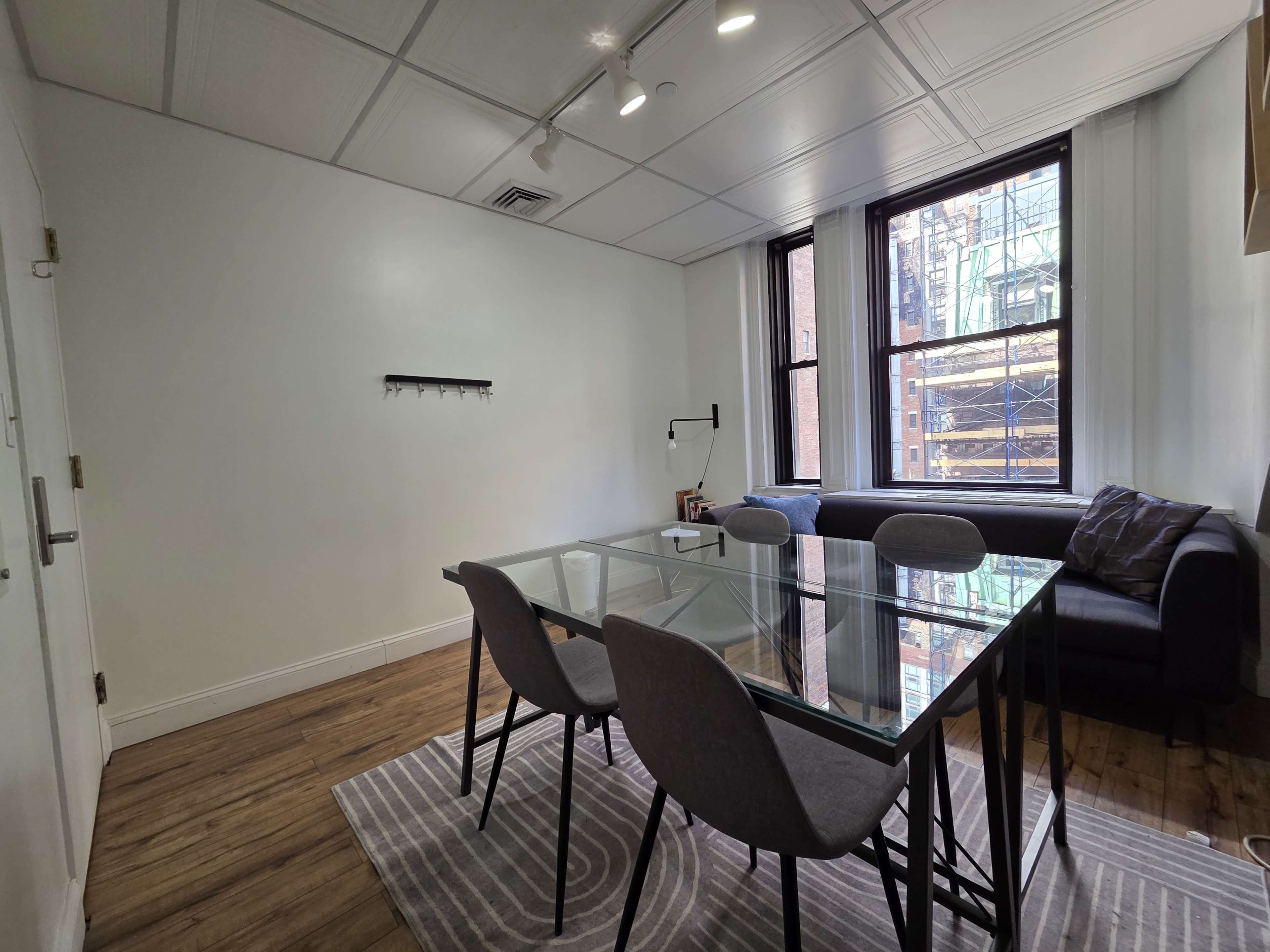 The image shows a modern room featuring a glass dining table surrounded by four gray chairs, a black sofa, and a window with a view of a construction site outside.