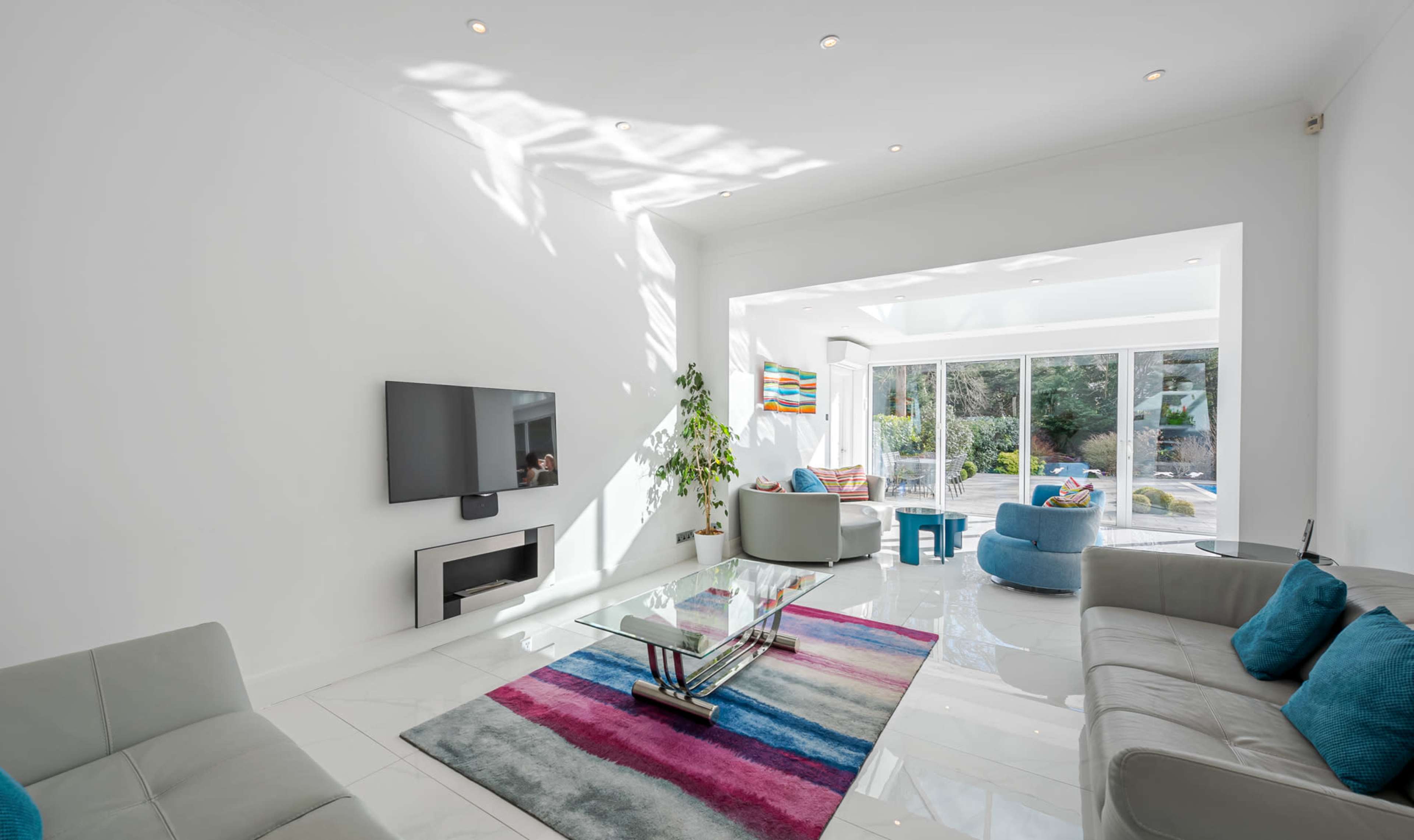 A modern living room featuring a large glass door that opens to an outdoor space, with light-colored furniture, a colorful rug, and a television mounted on the wall.