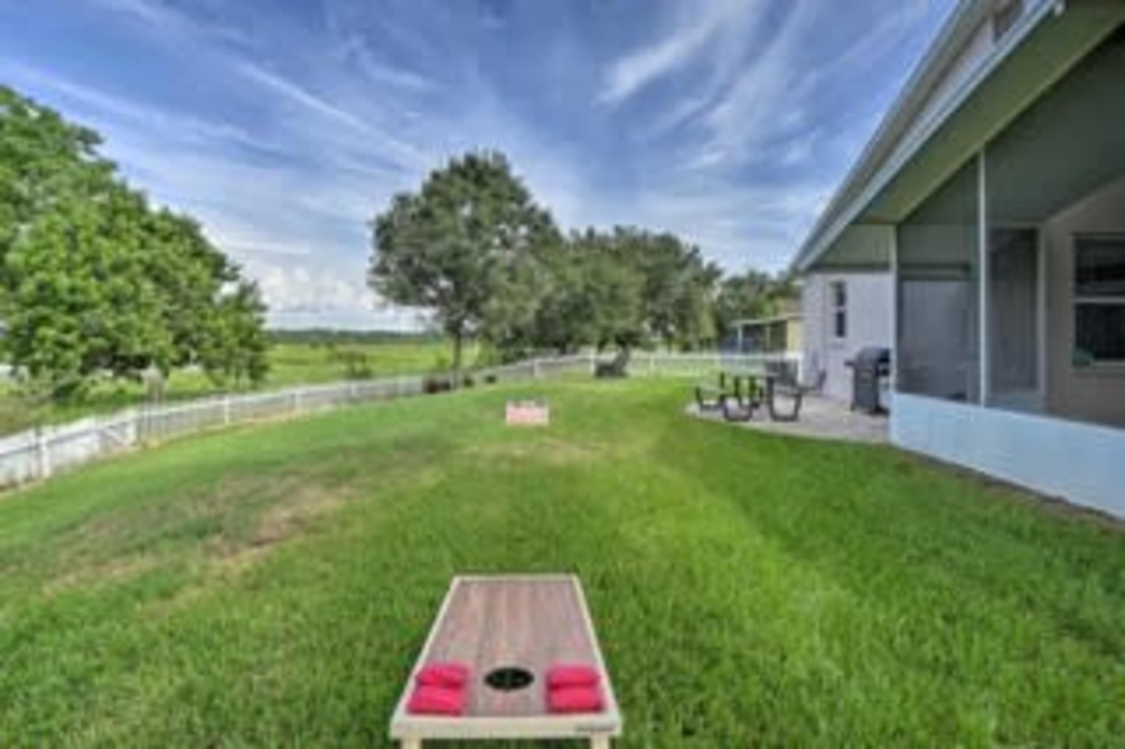 A grassy backyard features a cornhole game setup with bags, and a small picnic table nearby, against a backdrop of trees and a clear sky.