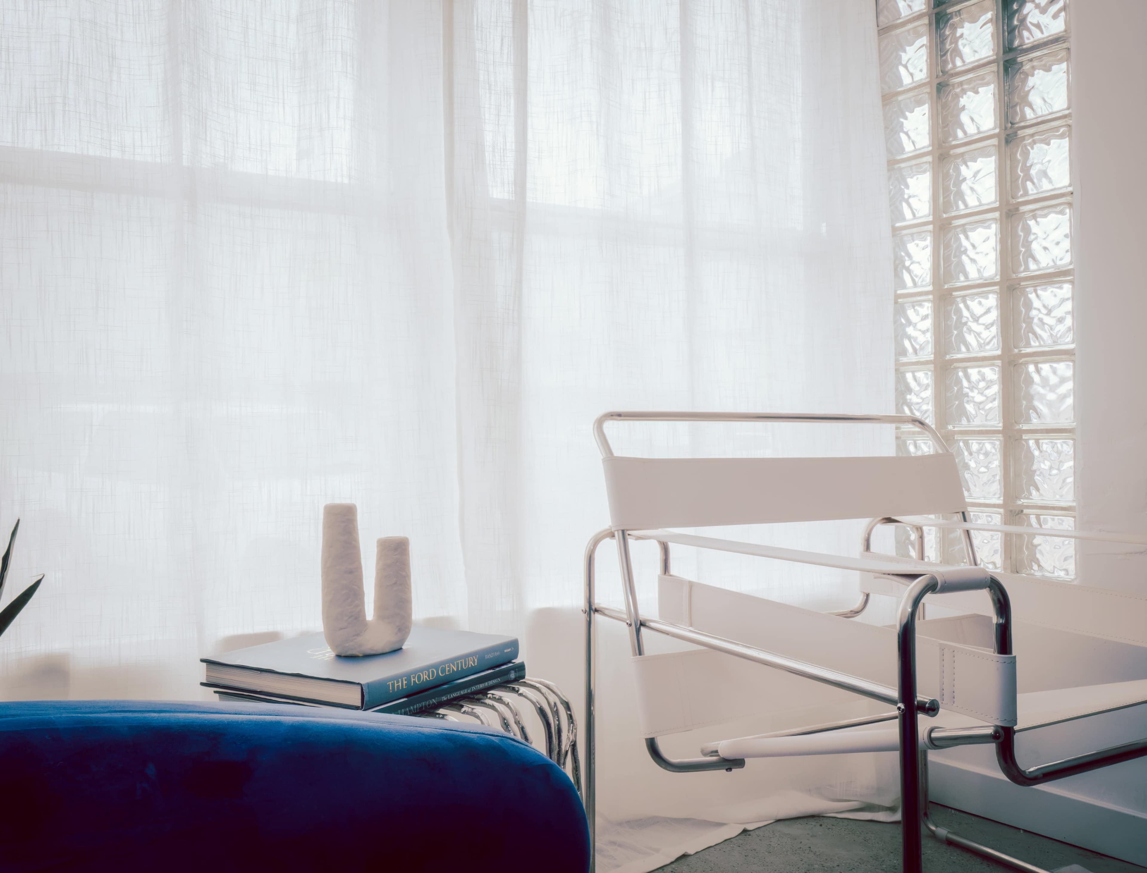 The image features a modern, minimalist room with a white chair, a small stack of books, and two decorative vases beside a large window with sheer curtains.