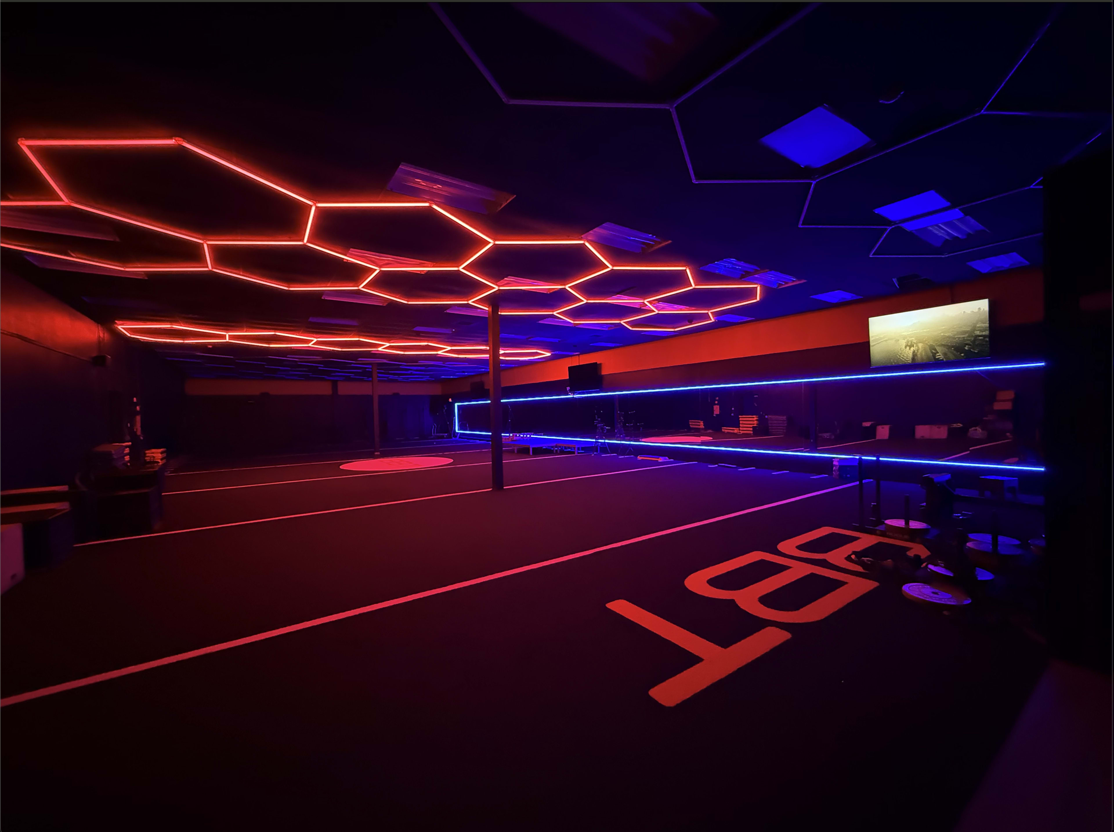 The image shows a modern indoor space with hexagonal neon lighting in red and blue, featuring a dark floor and a television screen on one wall.