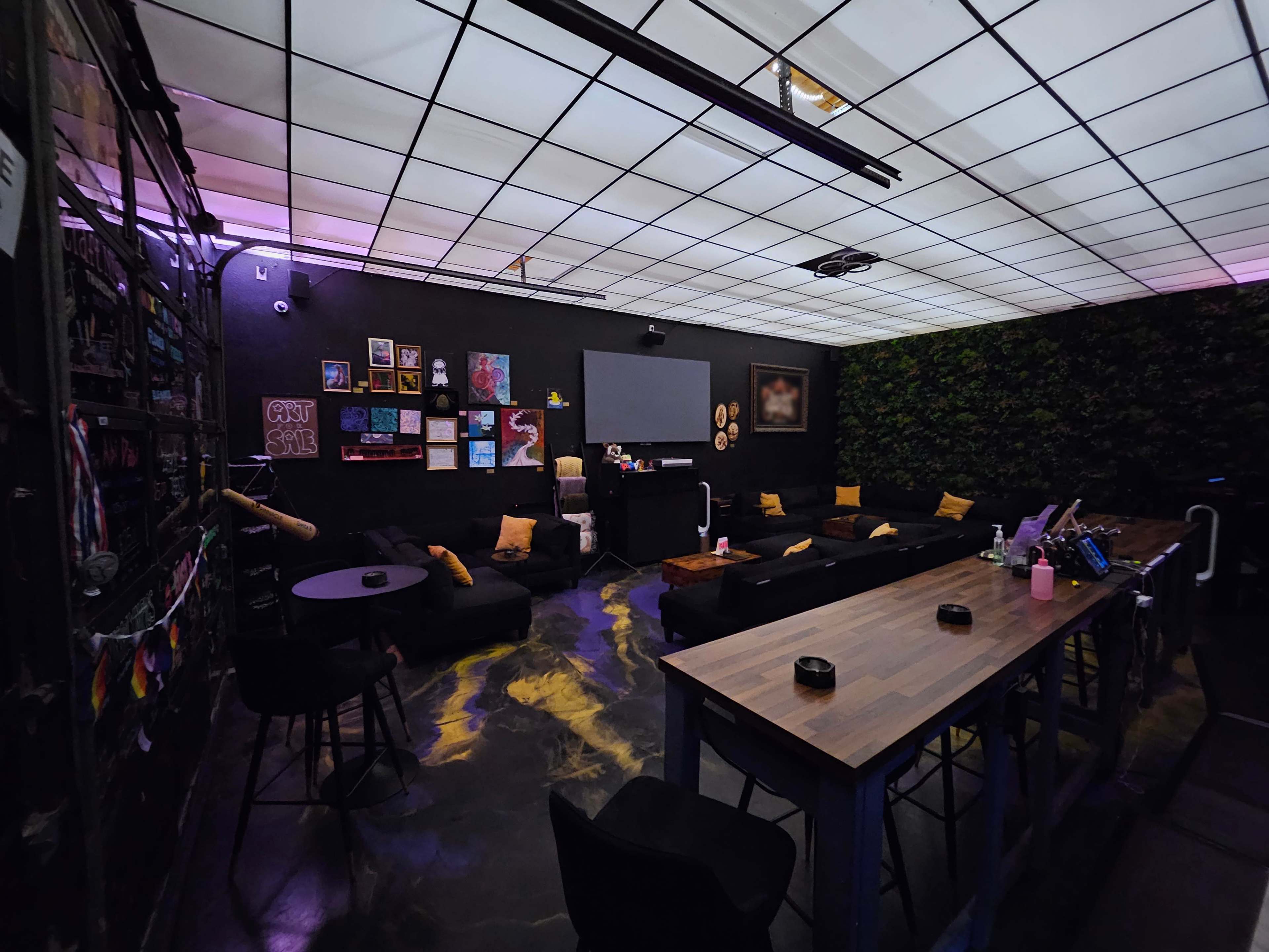 The image depicts a dimly lit lounge area with black sofas, a large screen, and a long table, surrounded by dark walls adorned with artwork.
