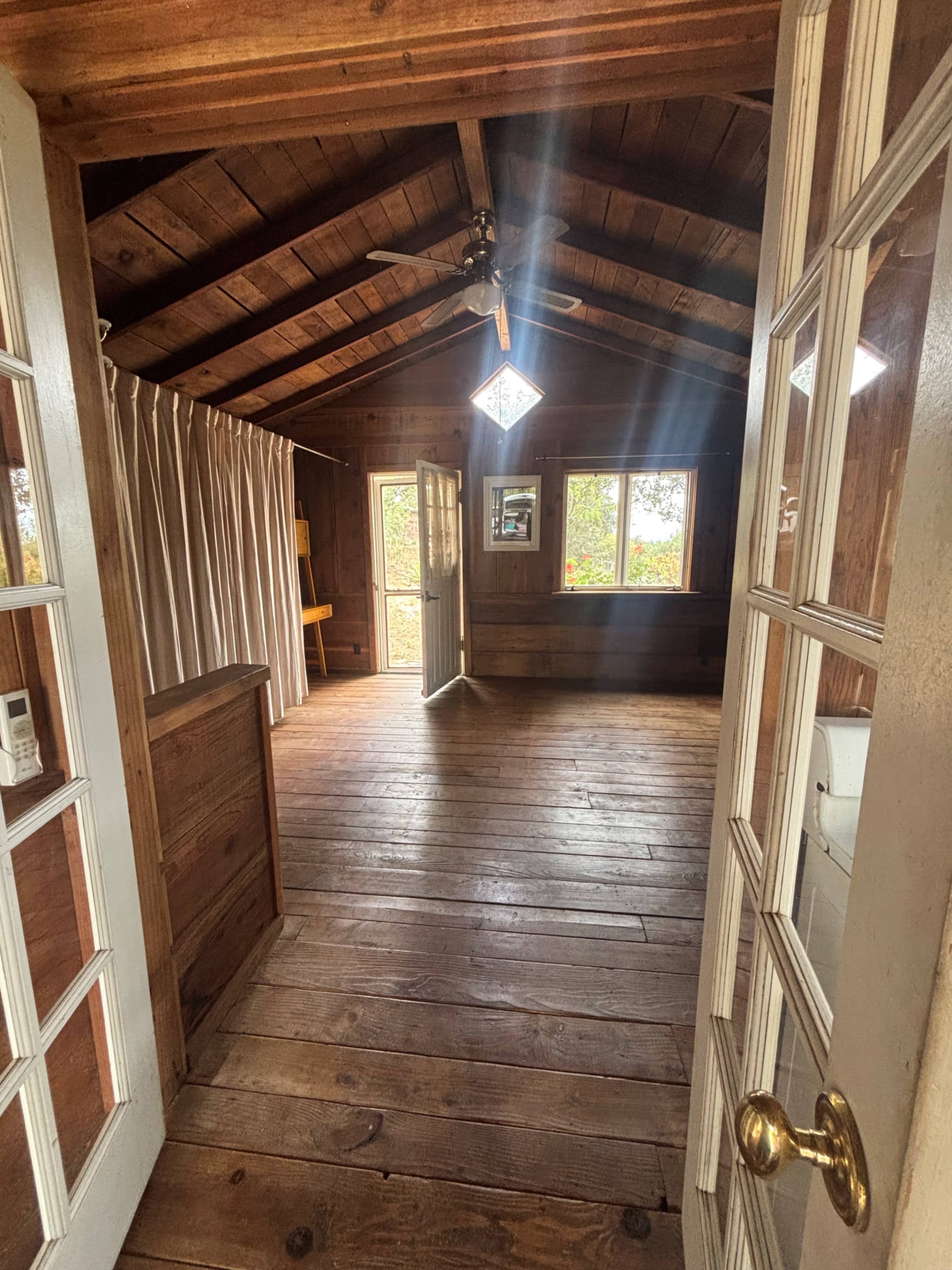 The image shows the interior of a wooden room with a sloped ceiling and large windows, featuring two open doors and wooden flooring.