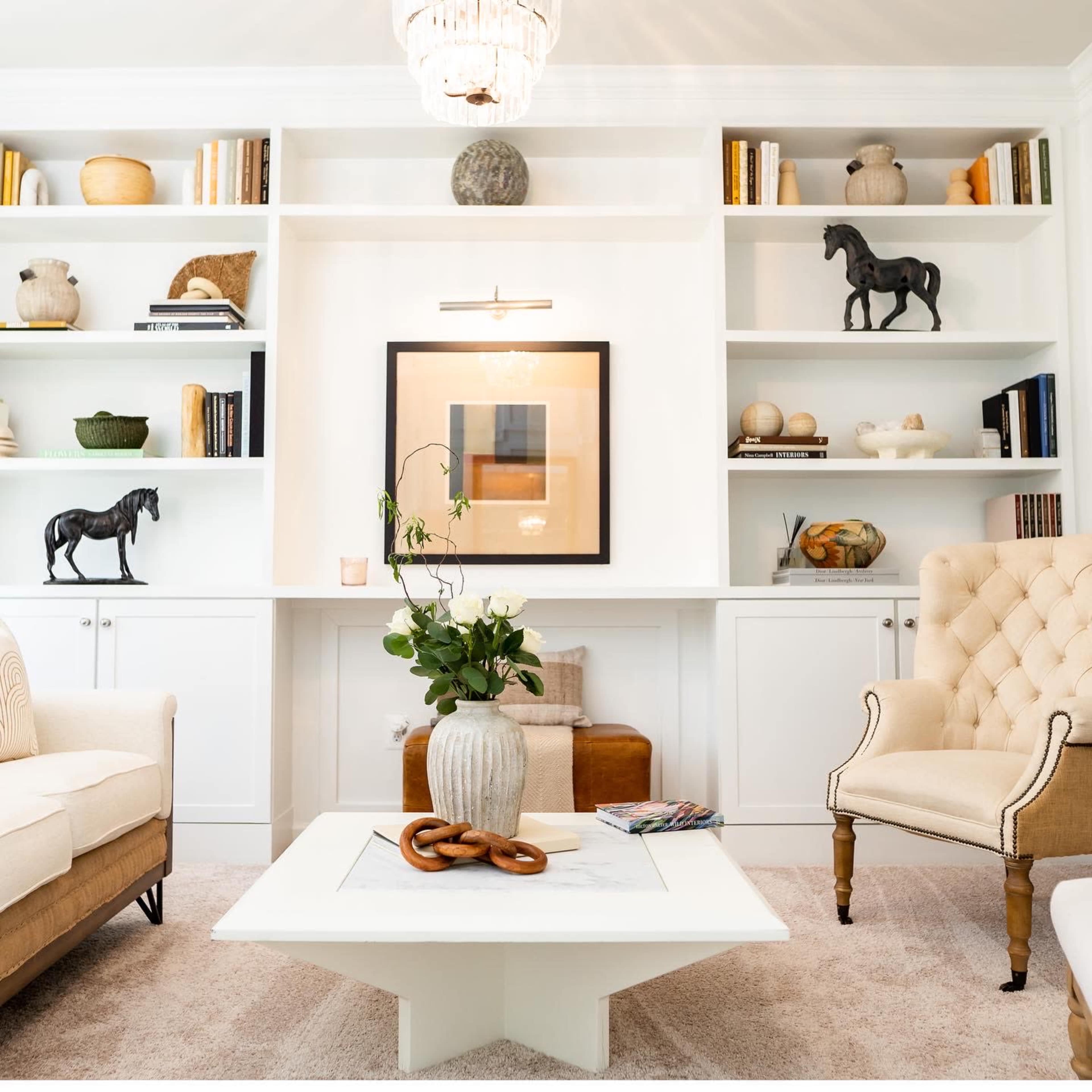 A bright living room features a light-colored sofa, a tufted armchair, a central white coffee table, and neatly arranged bookshelves filled with decorative objects and books.