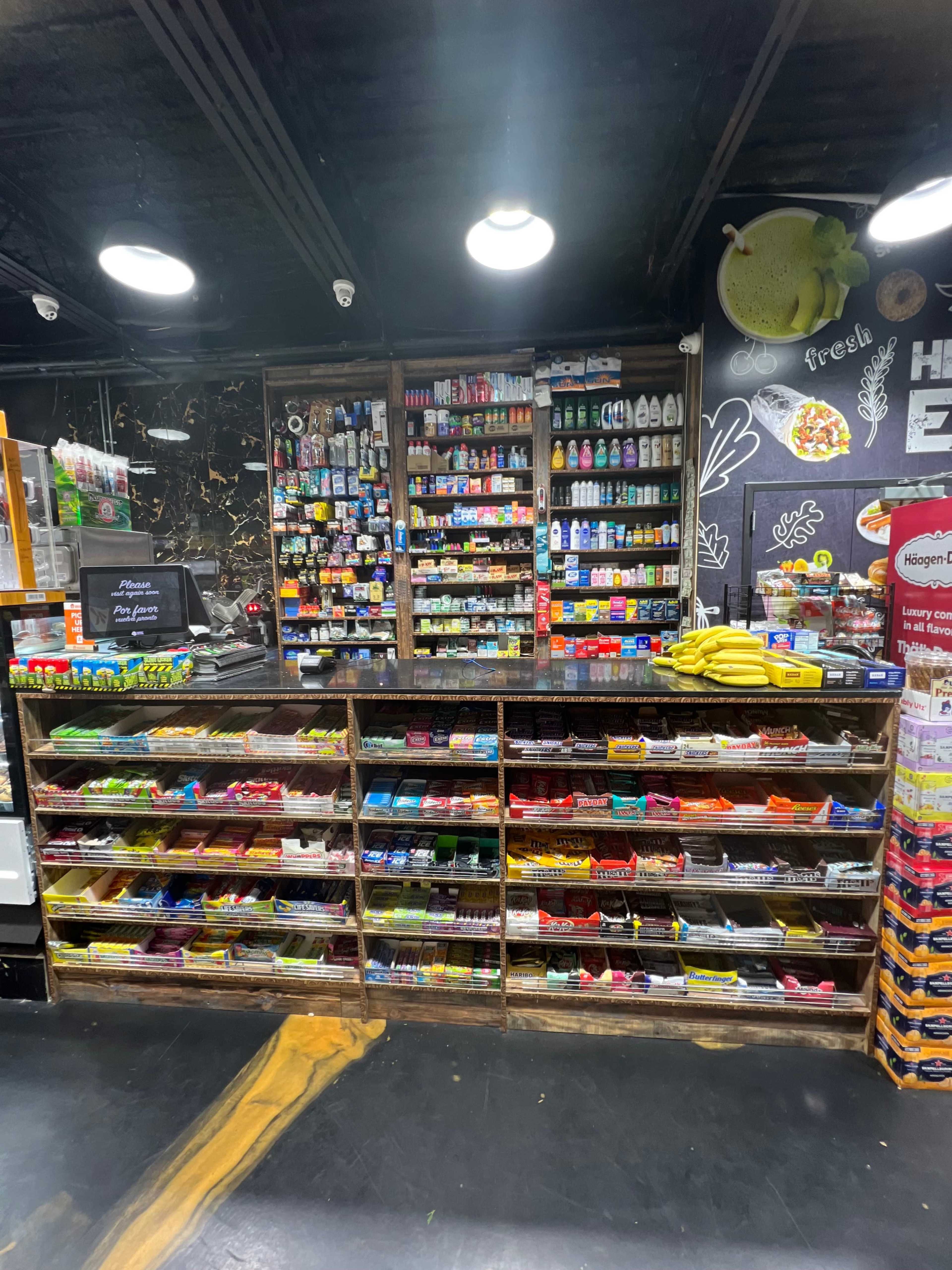 A cluttered convenience store counter displays a variety of snacks, drinks, and household items organized in wooden shelves.