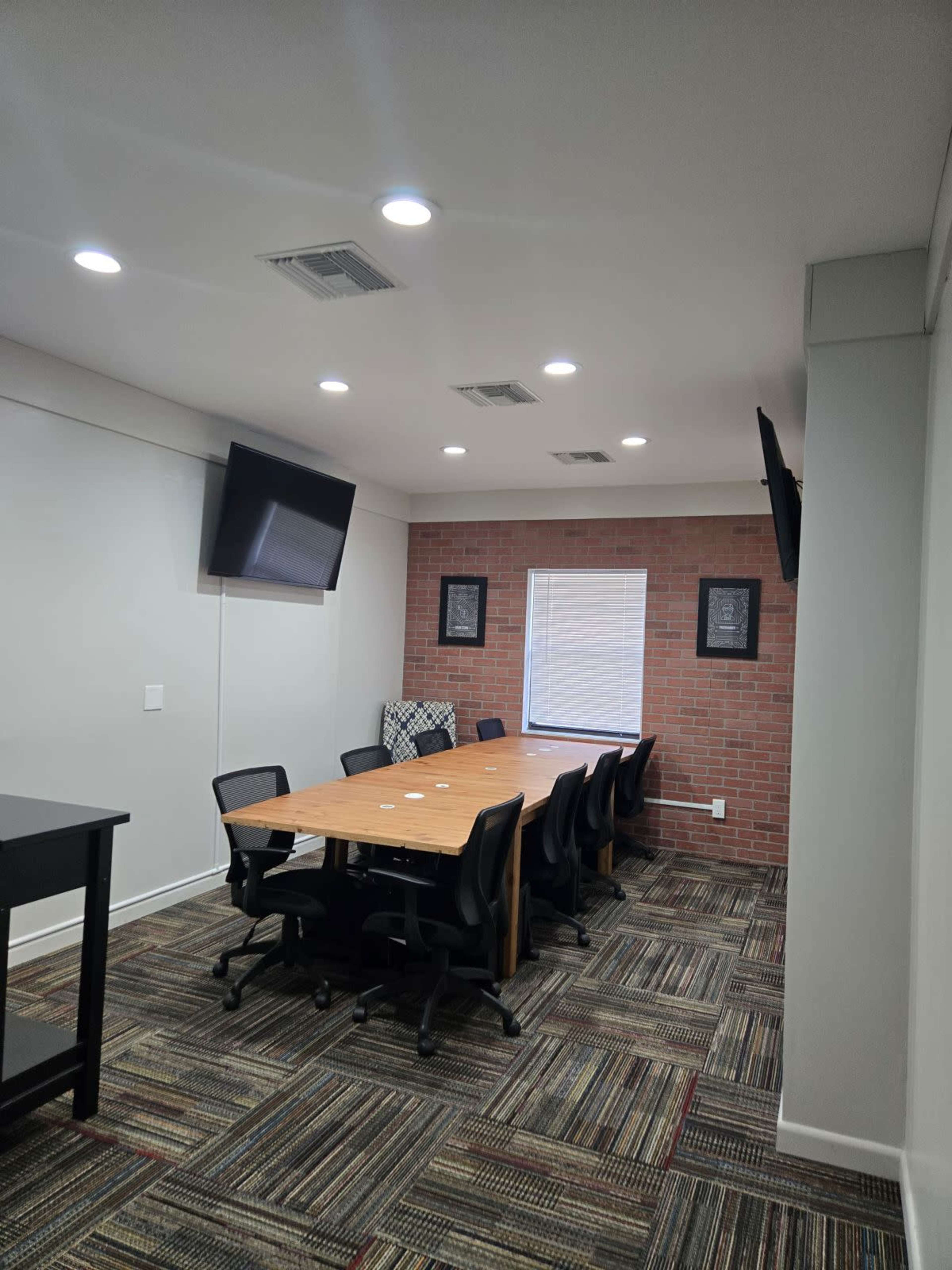 The image shows a conference room with a long wooden table surrounded by black office chairs, two wall-mounted TVs, and a window with a view of a brick wall.