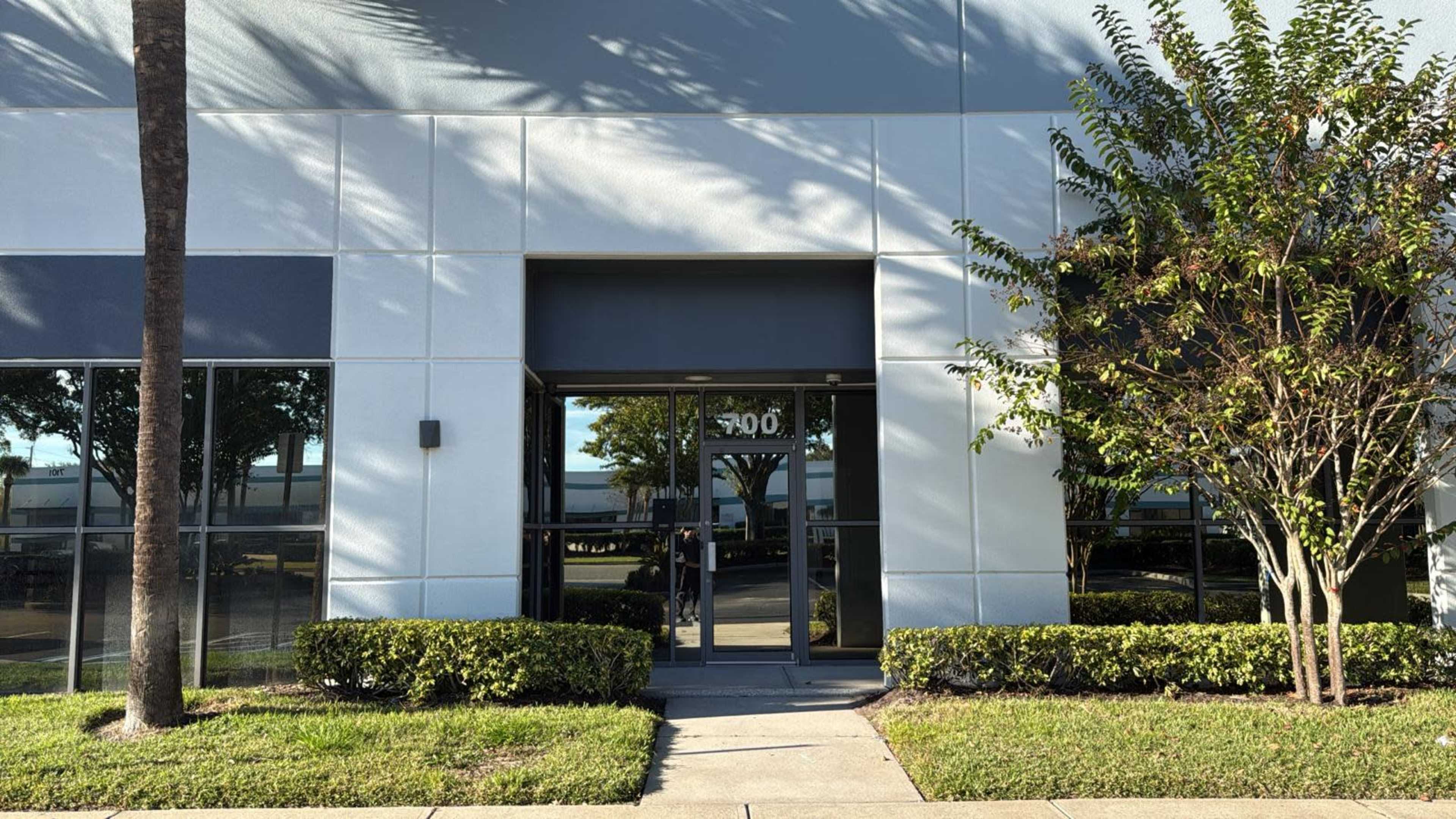 The image shows a modern commercial building entrance framed by neatly trimmed shrubs and palm trees.