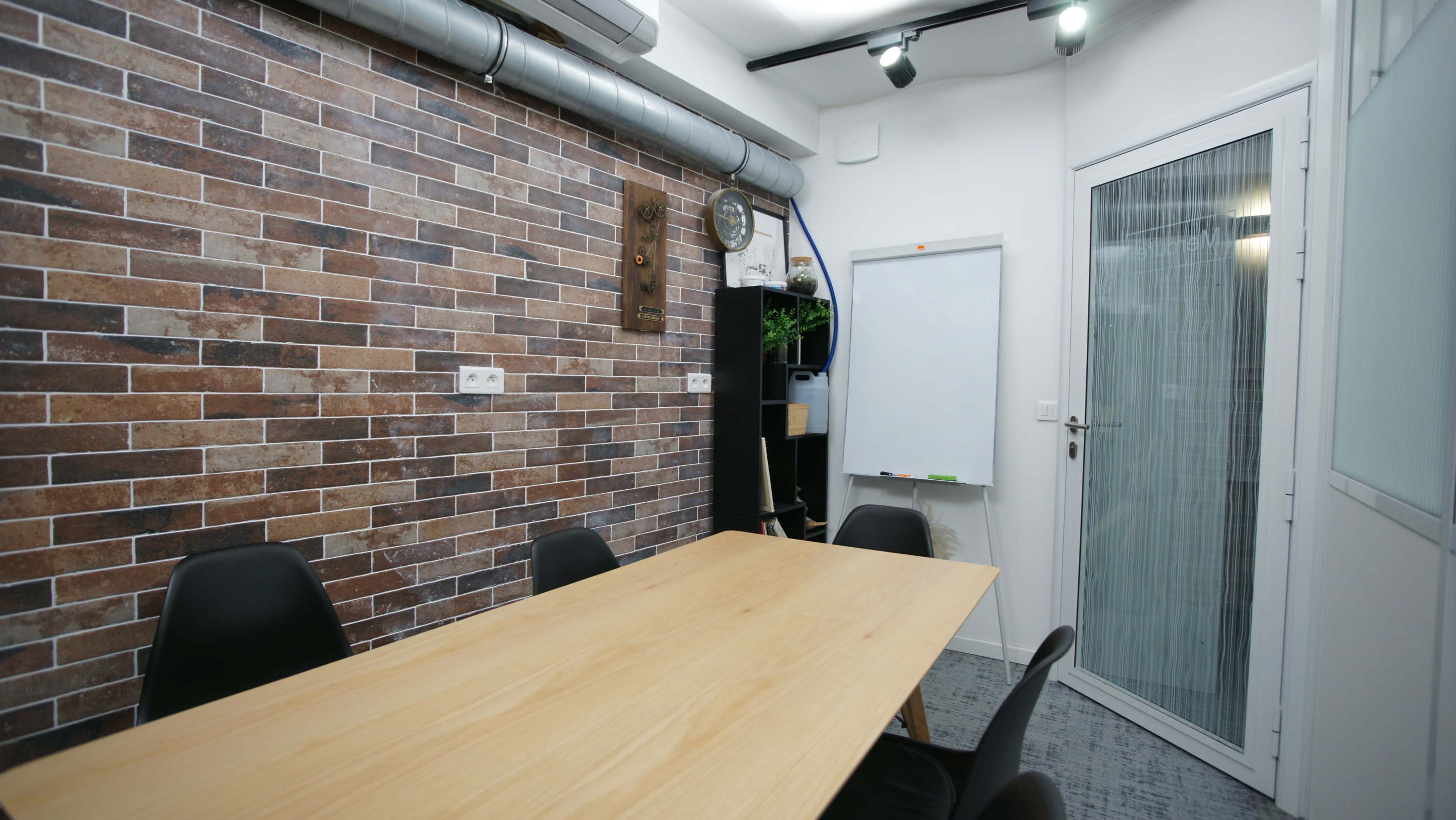 A small meeting room with a wooden table, black chairs, a brick-patterned wall, and a whiteboard on the side.