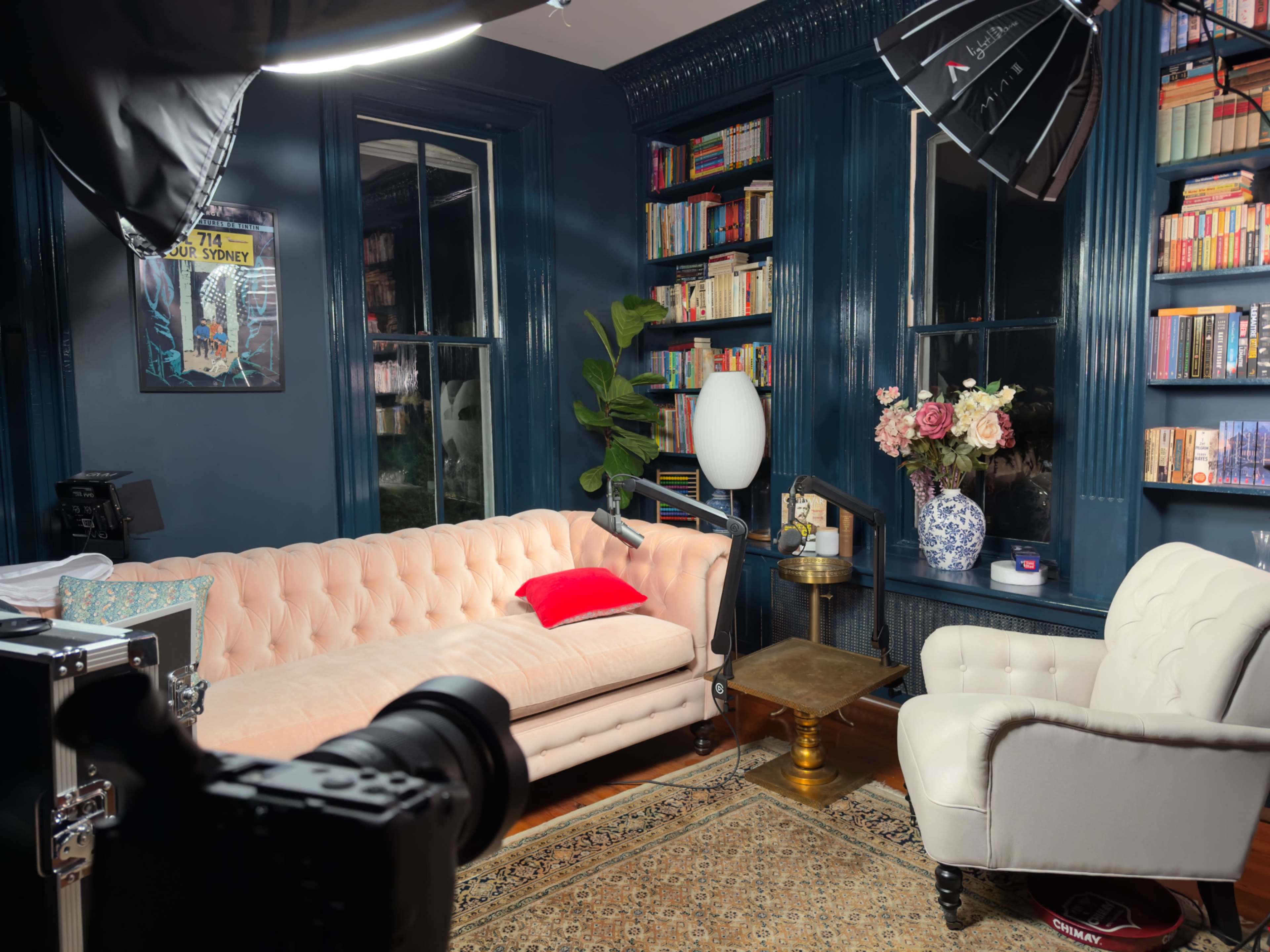 The image shows a cozy reading nook with a light pink tufted couch, a white armchair, a gold side table, and bookshelves filled with books, all set against dark blue walls.
