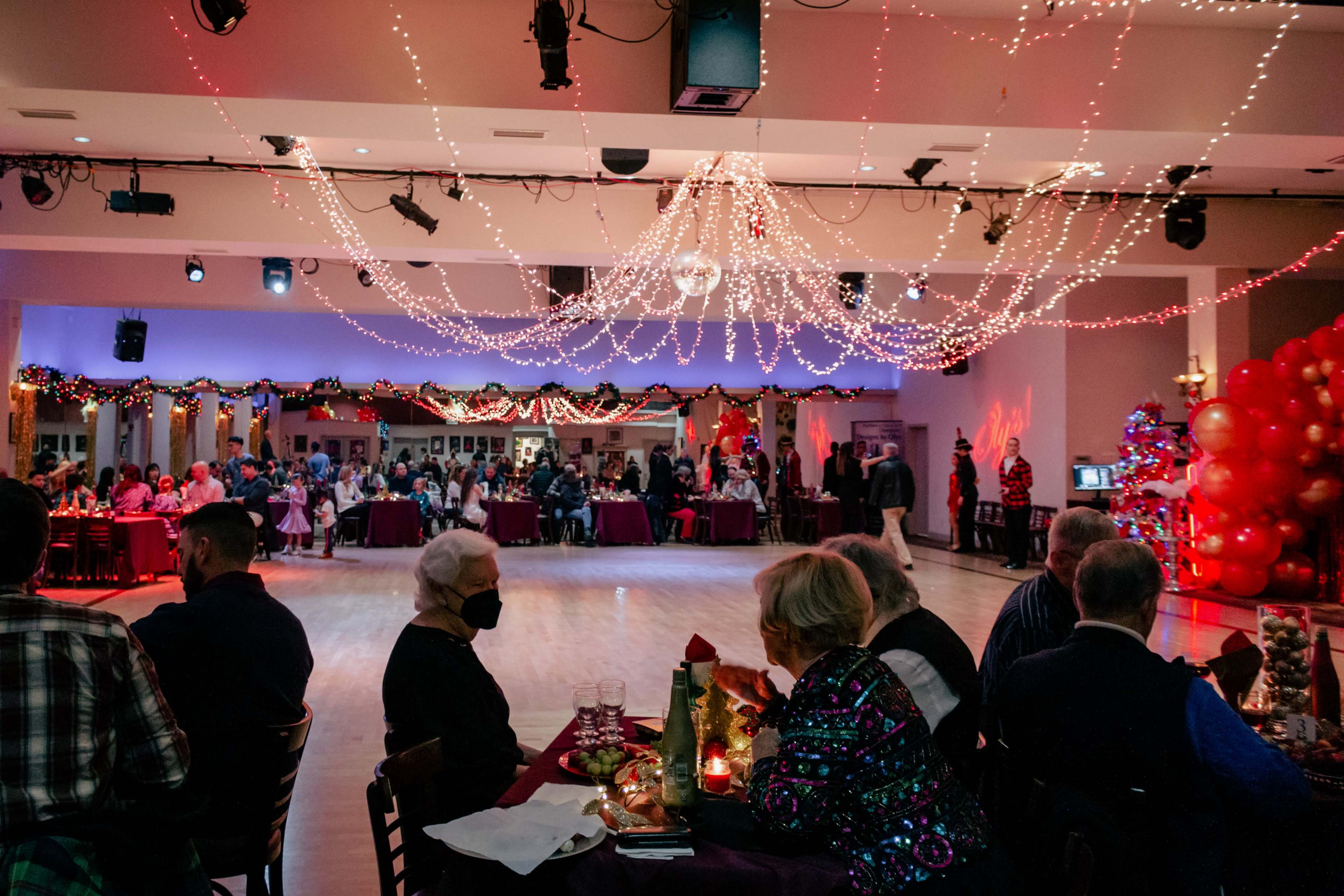 A festive dining hall is decorated with string lights and a Christmas tree, filled with guests seated at tables and enjoying a holiday celebration.