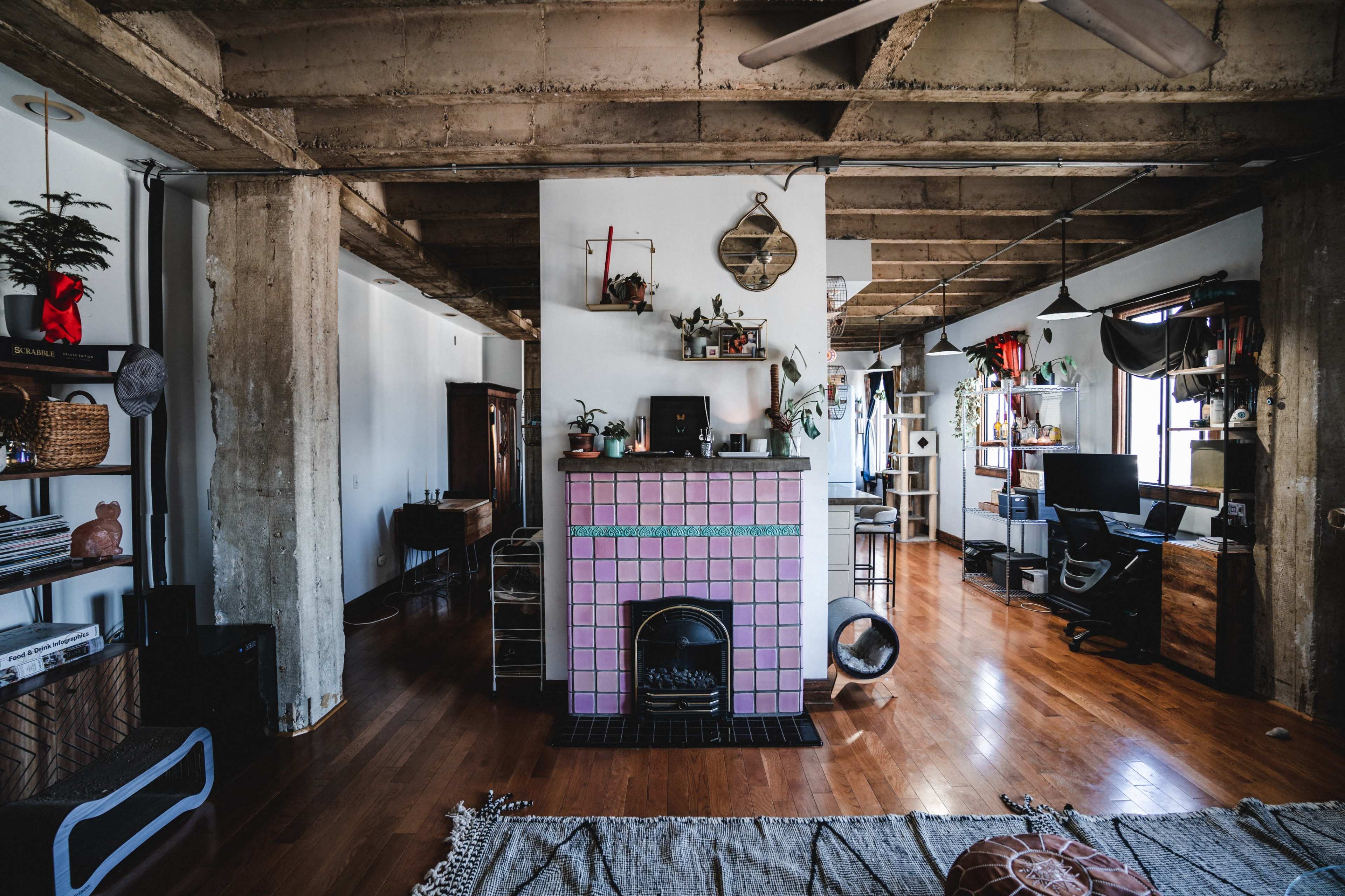 The image shows a spacious living area featuring a pink-tiled fireplace, wooden flooring, and various shelves filled with decorative items.