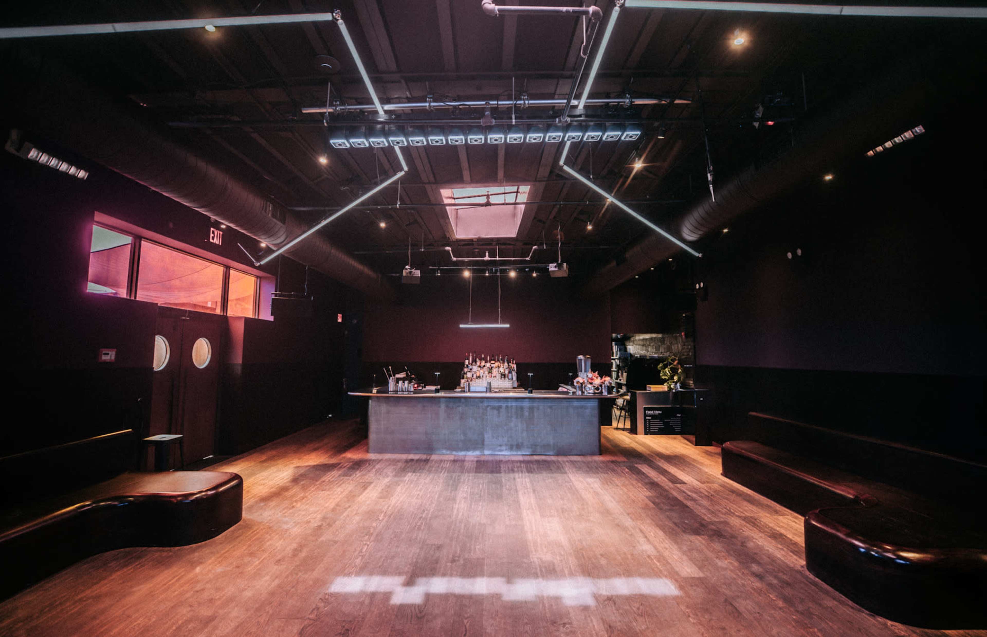 The image shows a minimalistic bar interior with a wooden floor, a long bar counter at the back, and seating along the walls.