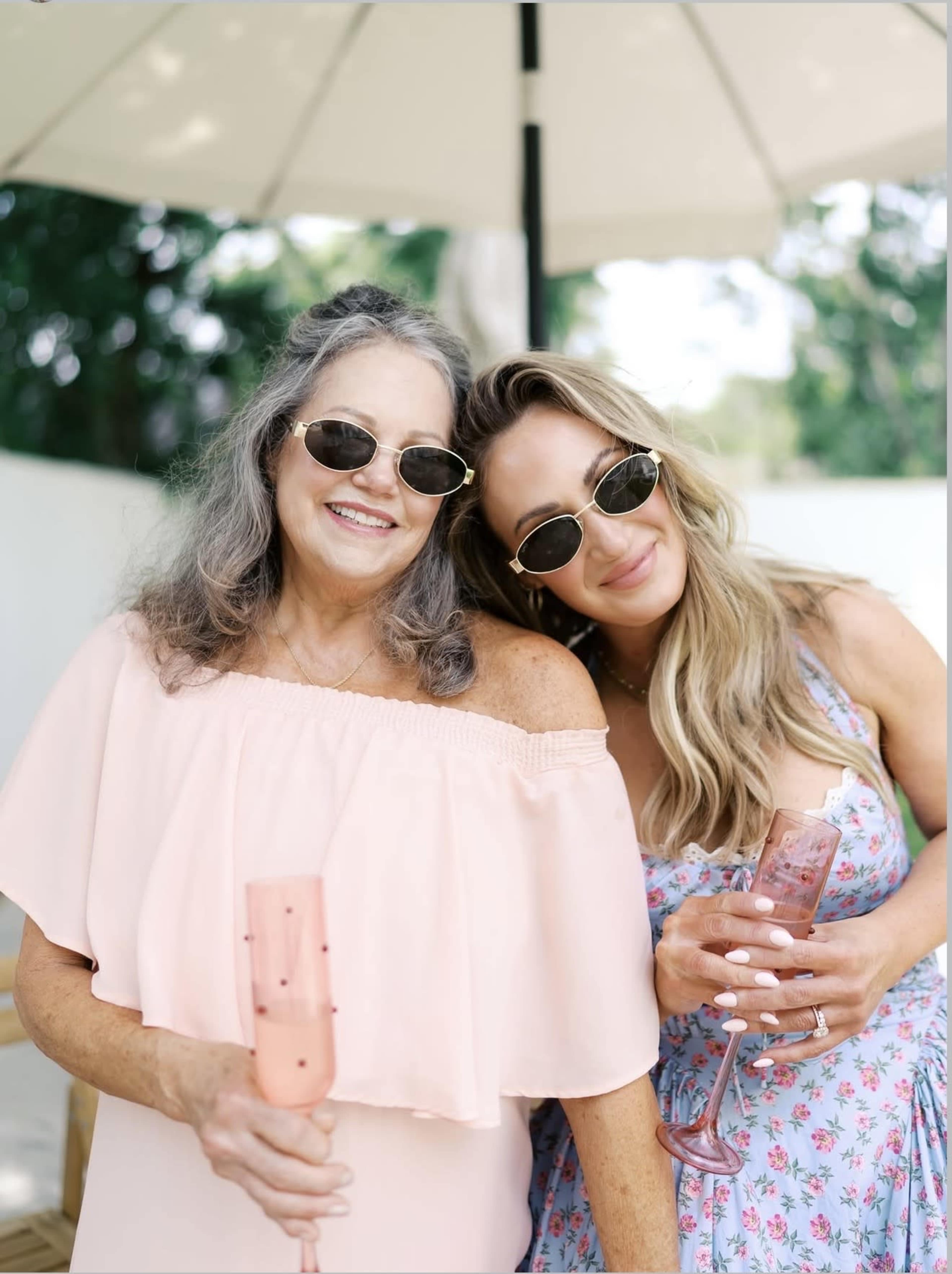 Two women, one with long hair and the other with gray hair, are smiling while holding champagne glasses under a large umbrella.