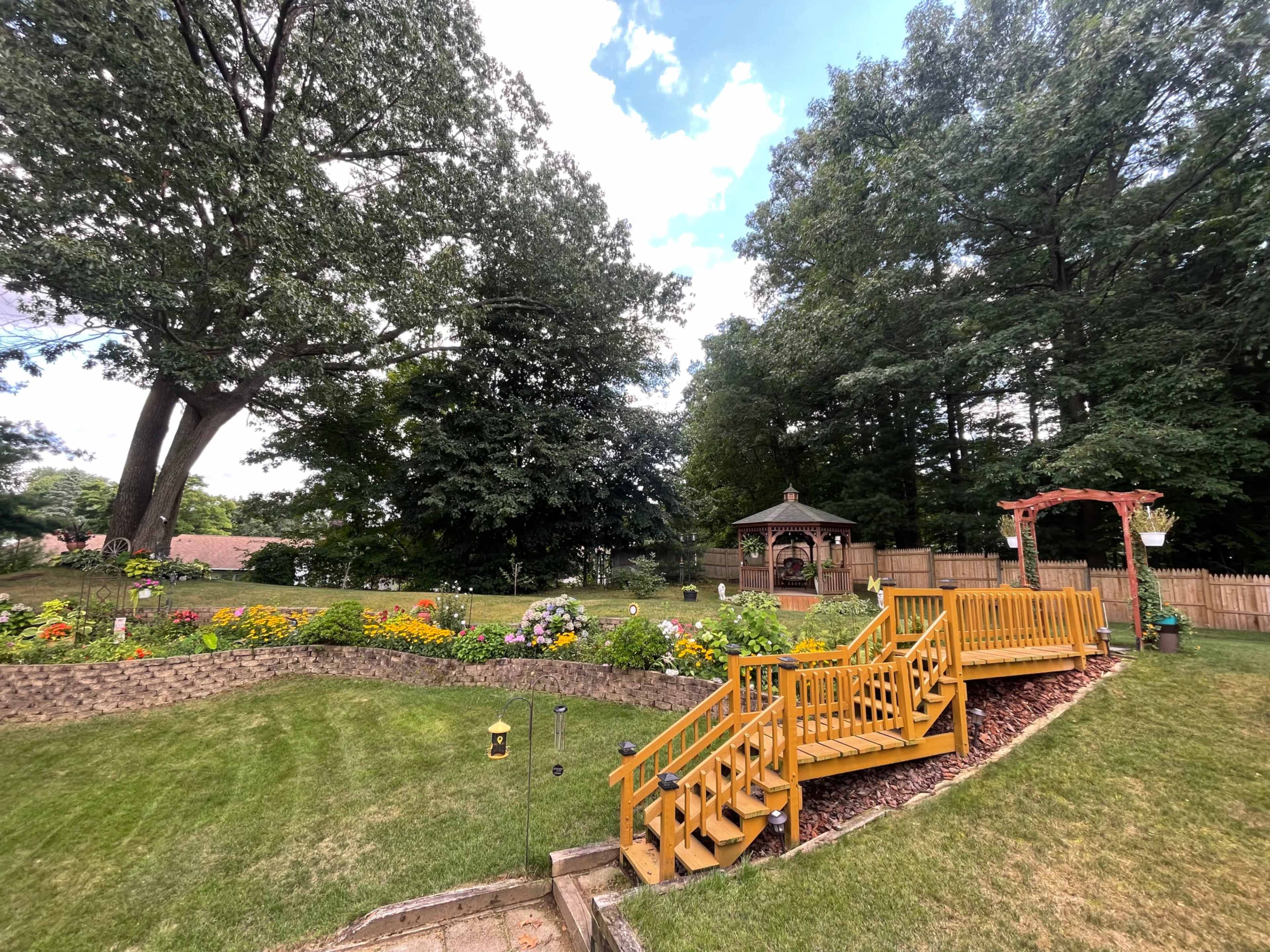 The image shows a landscaped backyard with colorful flower beds, a wooden deck with stairs, and a gazebo nestled among trees.