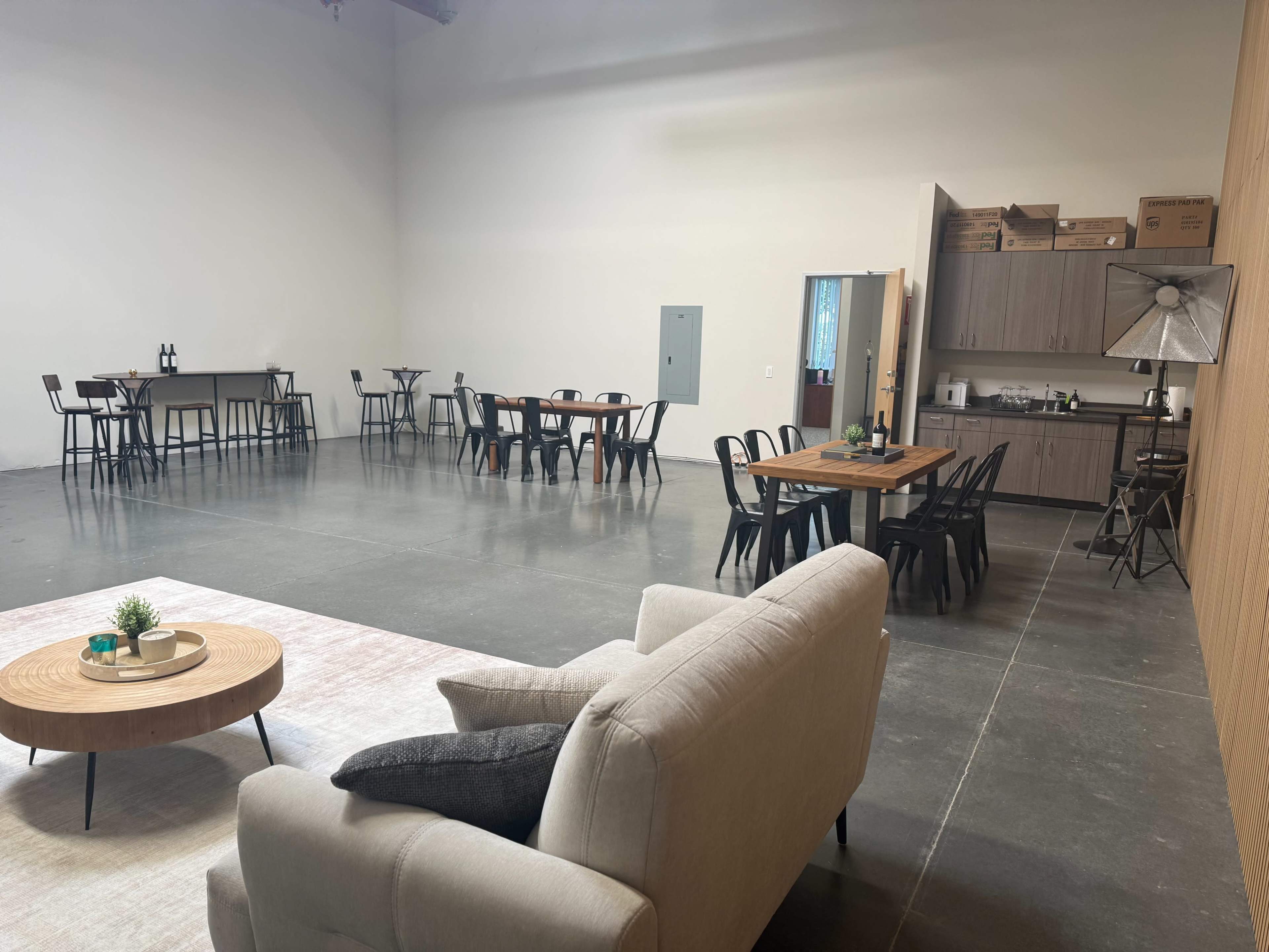The image shows a spacious, modern room arranged with several tables and chairs, along with a small kitchenette area and seating in one corner.