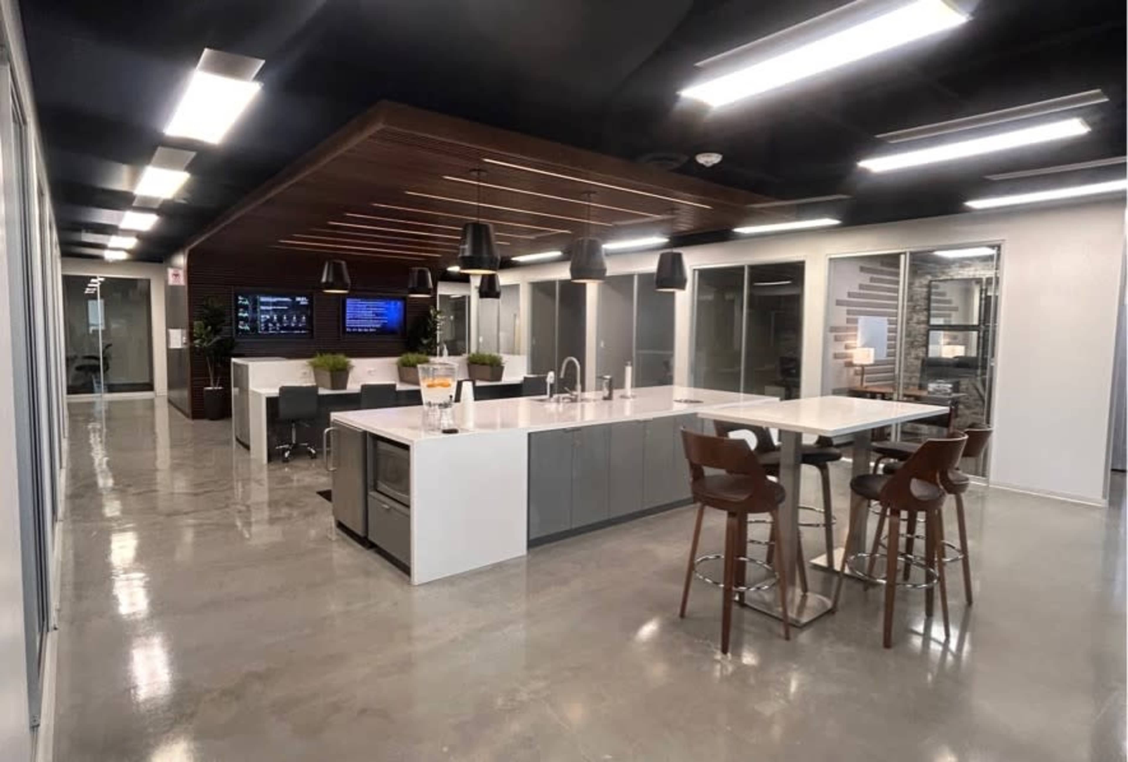 The image shows a modern office space featuring a central kitchen island, seating area, and large windows.