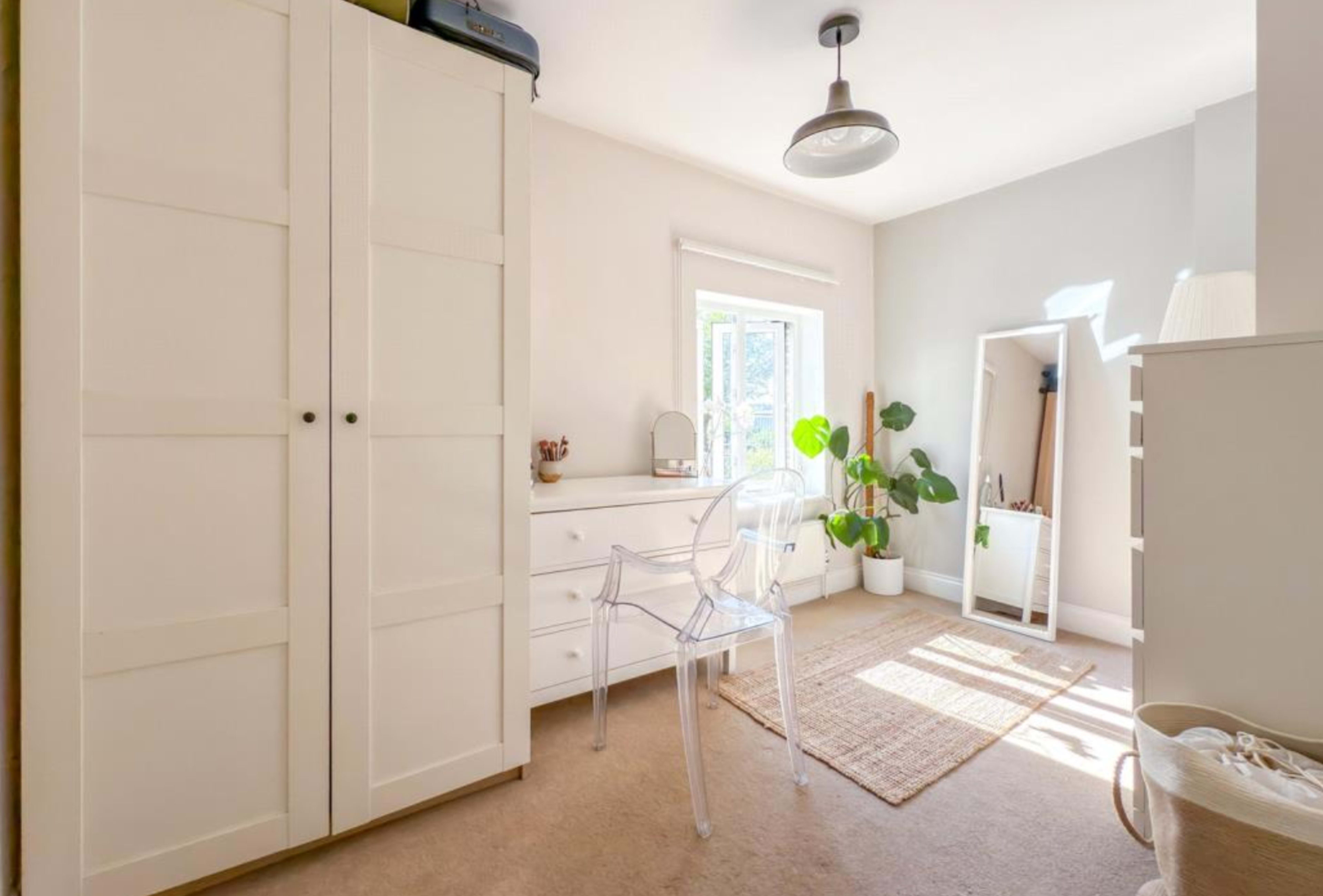 The image depicts a bright bedroom with light-colored walls, a clear chair, a dresser with drawers, a mirror, and a potted plant in the corner.