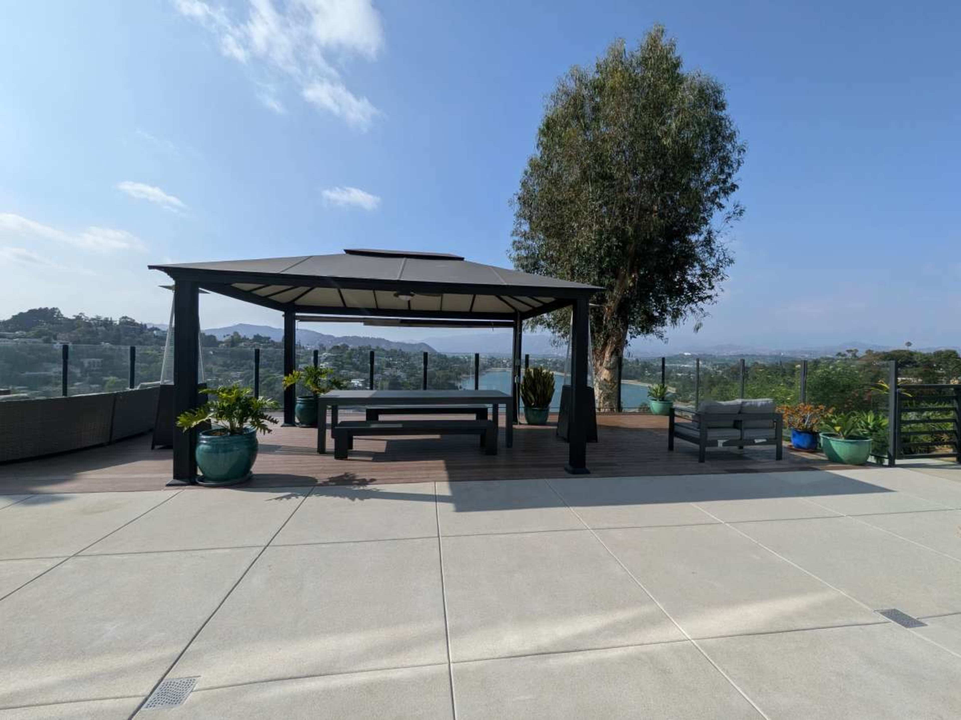 The image shows a open gazebo with seating surrounded by potted plants, overlooking a scenic landscape with distant hills and a clear sky.