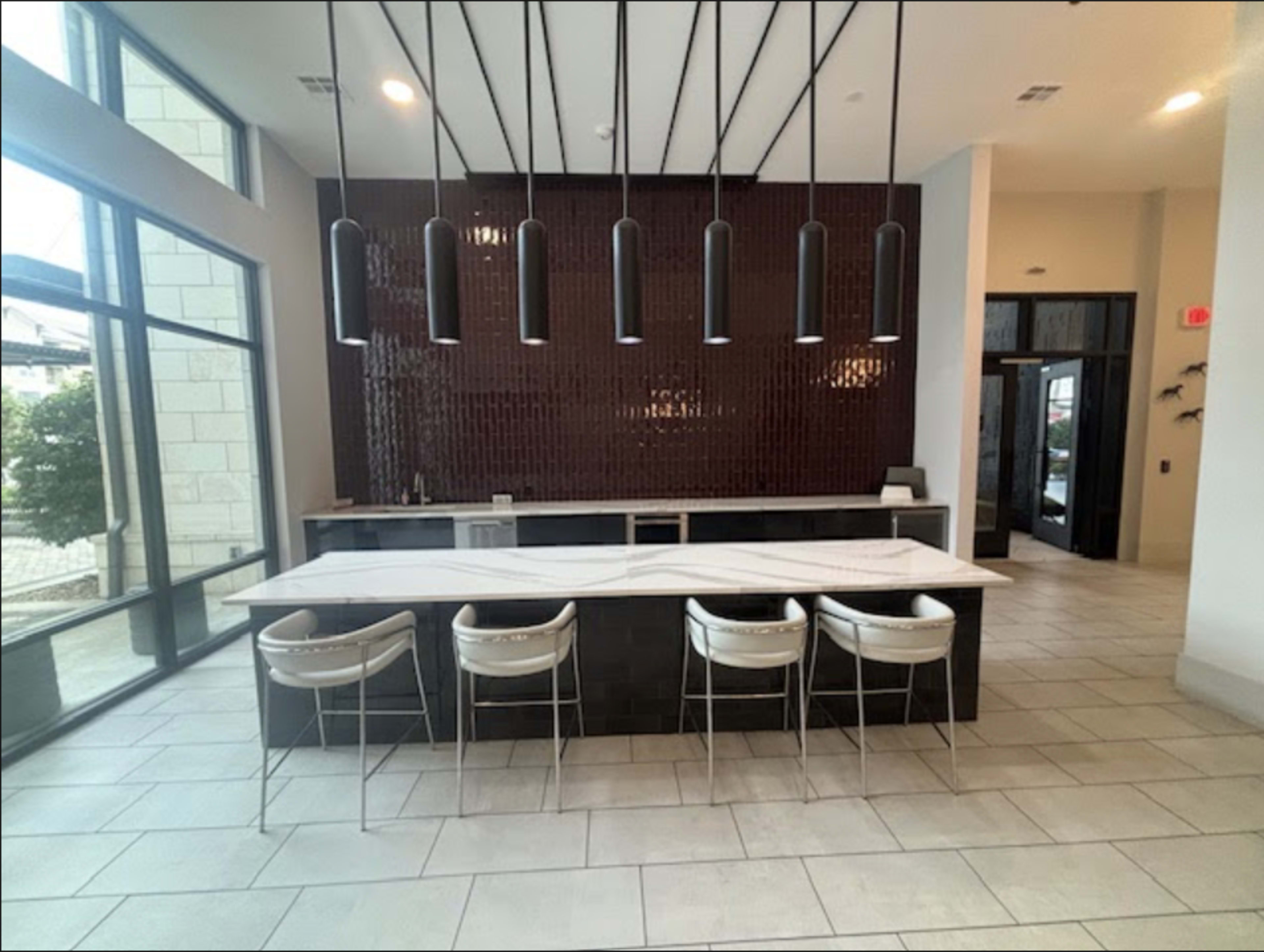The image shows a modern reception area featuring a long marble counter with four white stools and a dark tiled wall in the background, illuminated by six pendant lights.