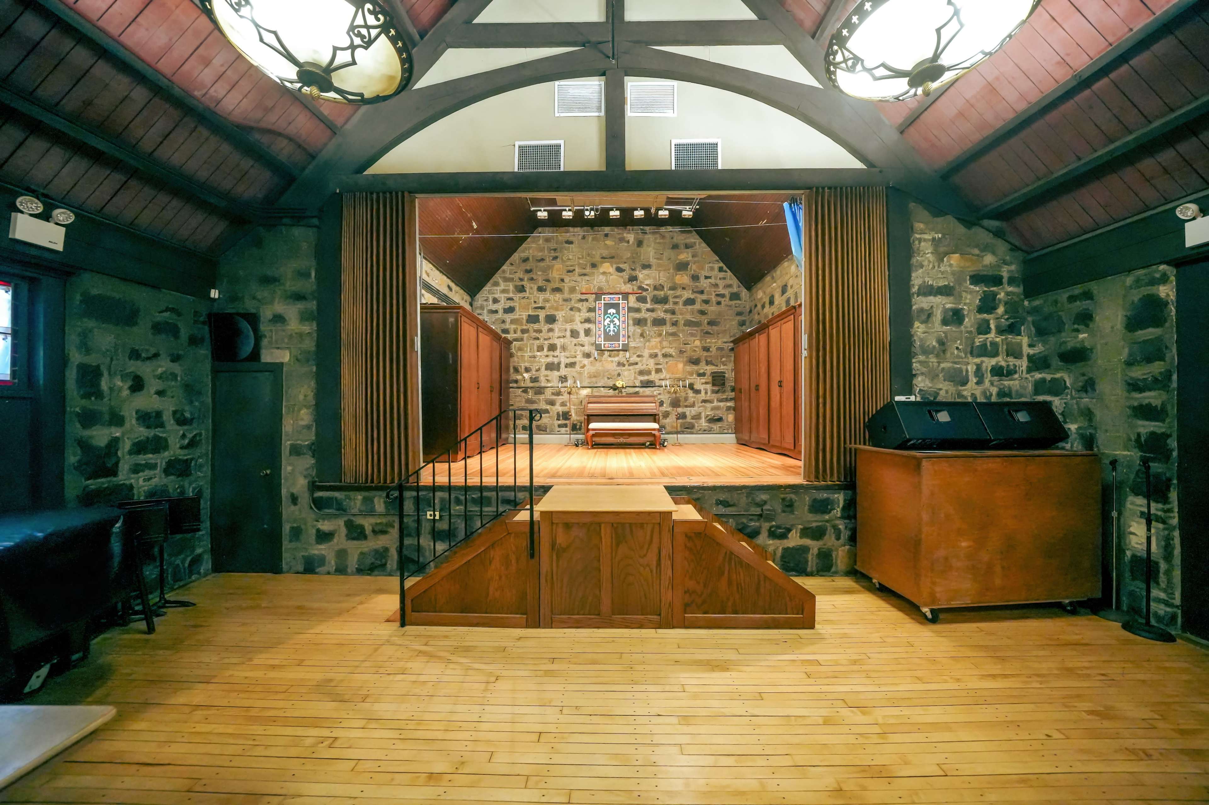 The interior of a stone-walled performance space with wooden flooring, featuring a stage at the front and a staircase leading up to it.