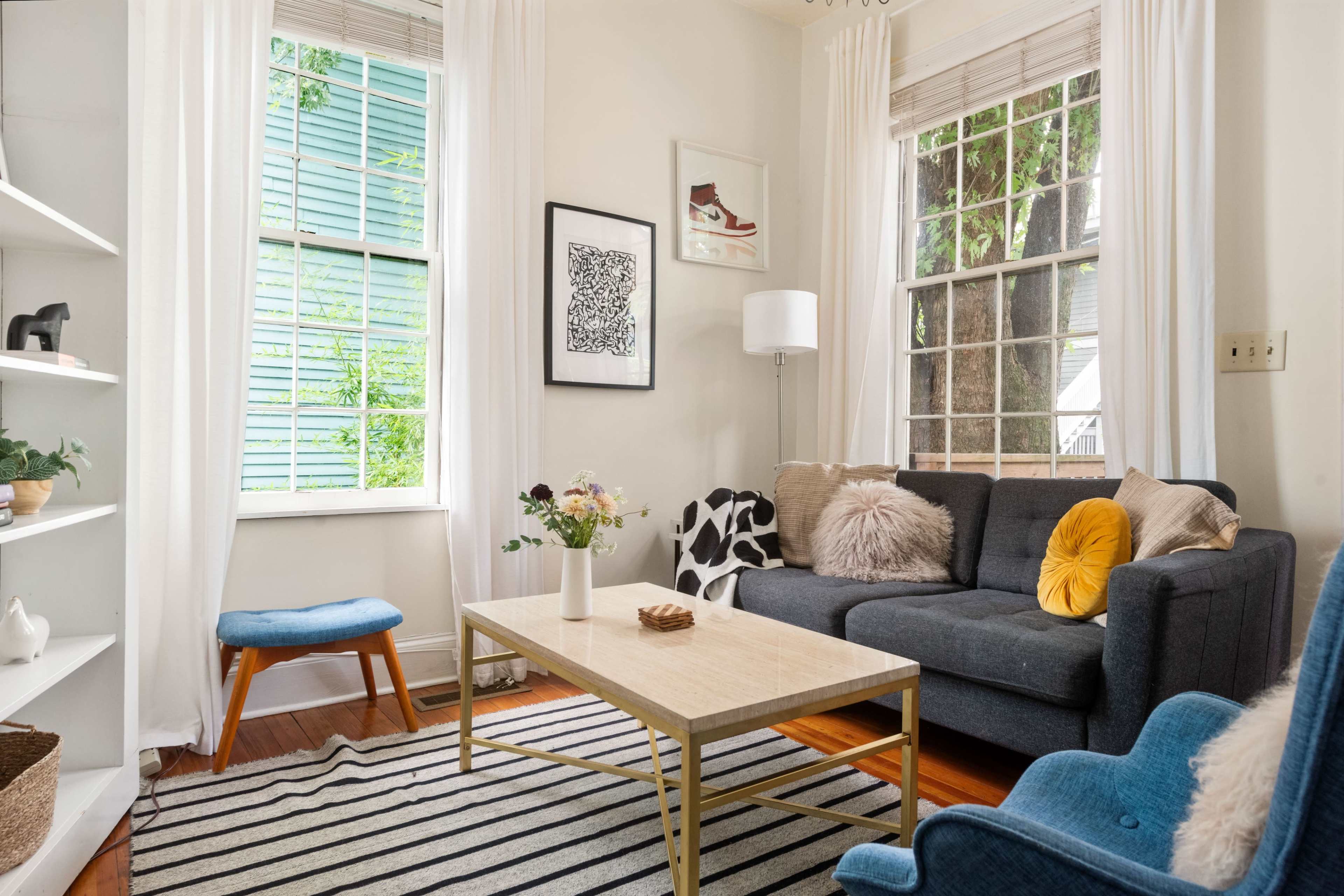 A cozy living room features a gray sofa, a wooden coffee table, and two chairs, with large windows providing natural light and a view of greenery outside.
