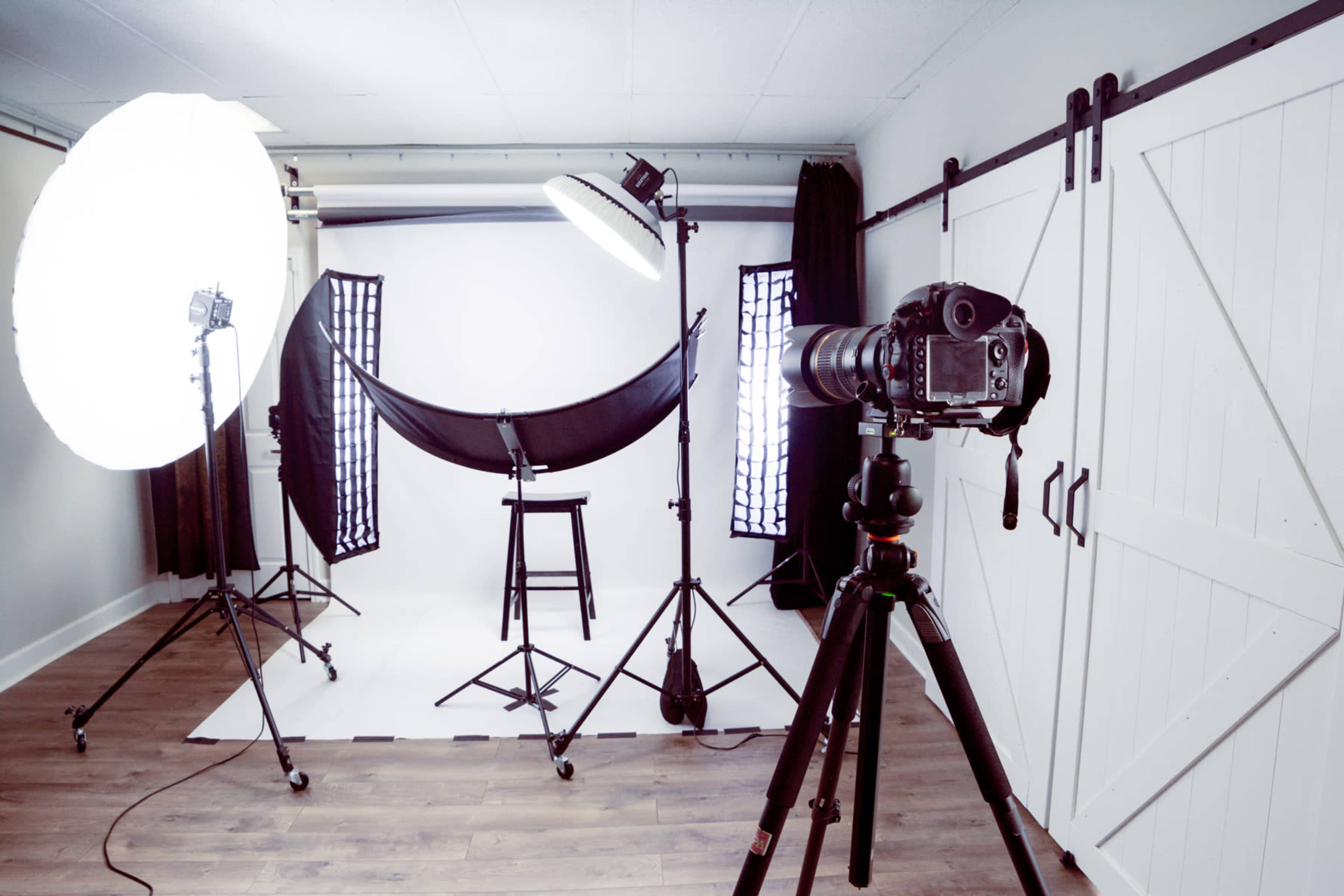 A photography studio setup with large softbox lights, a backdrop on stands, a stool, and a camera mounted on a tripod.