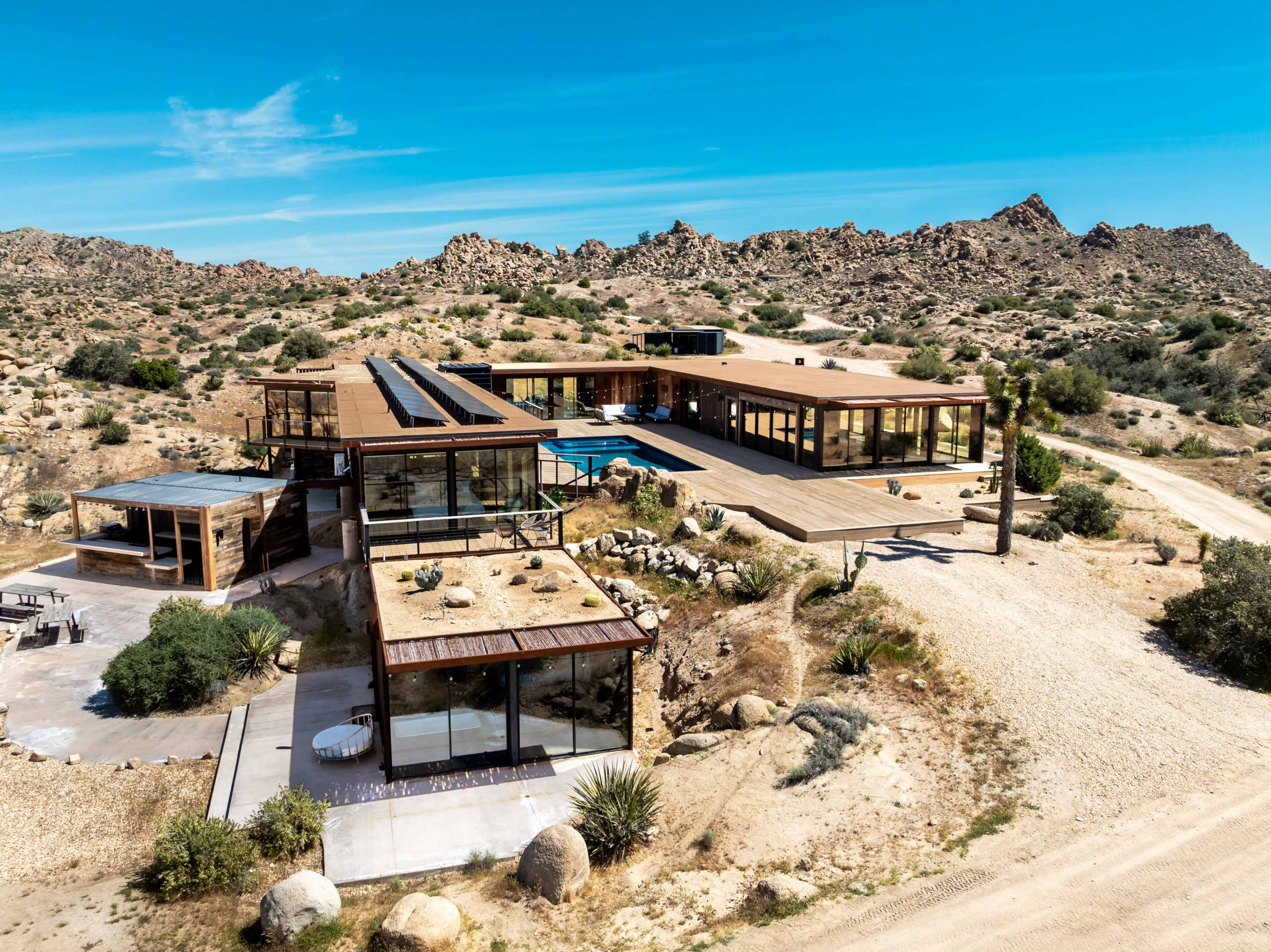 The image shows a modern architectural home situated in a rocky desert landscape, featuring large glass windows and multiple outdoor terraces.