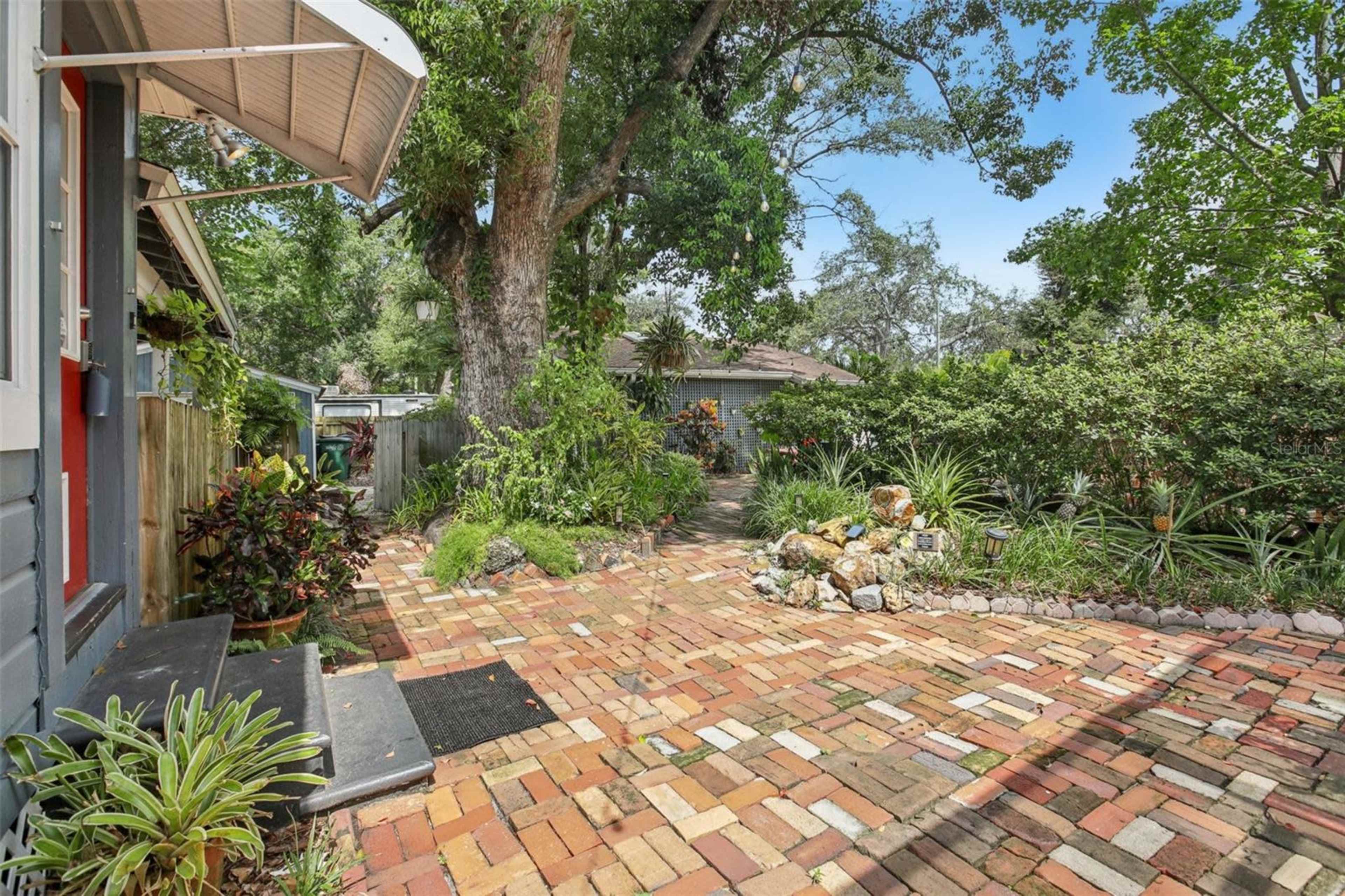 The image shows a brick pathway leading to a house, surrounded by various trees and plants.