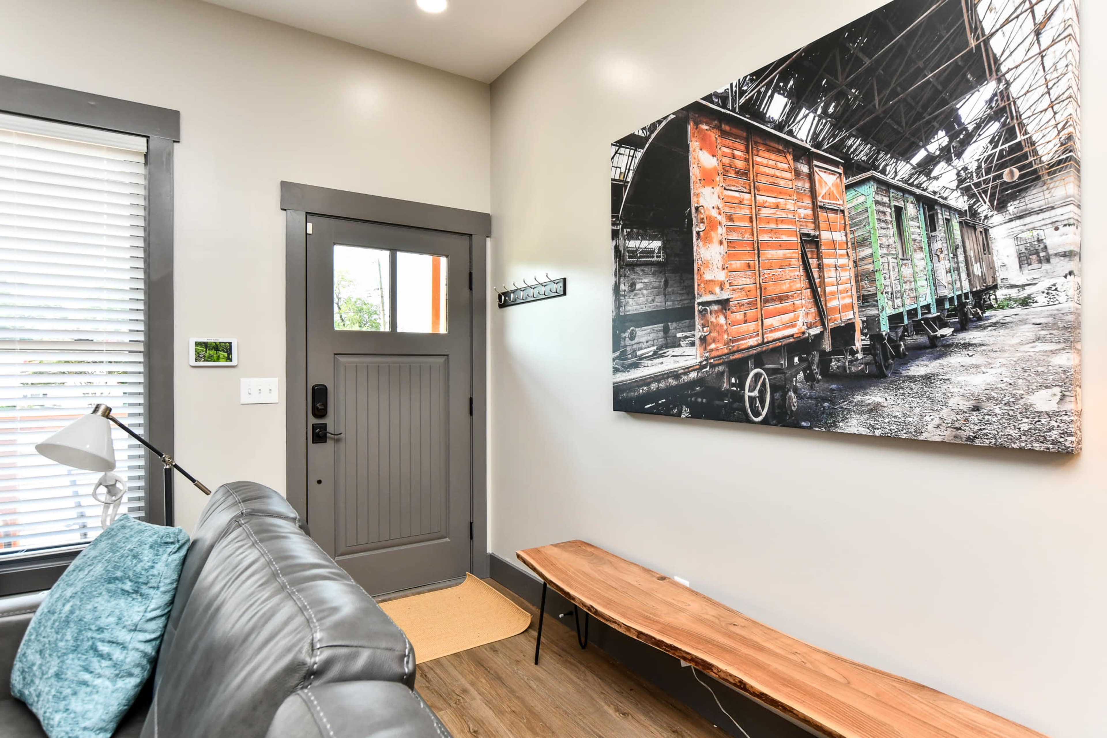 An interior entrance area with a door, a bench made of wood, a gray couch, and a large wall-mounted photograph of colorful train cars.