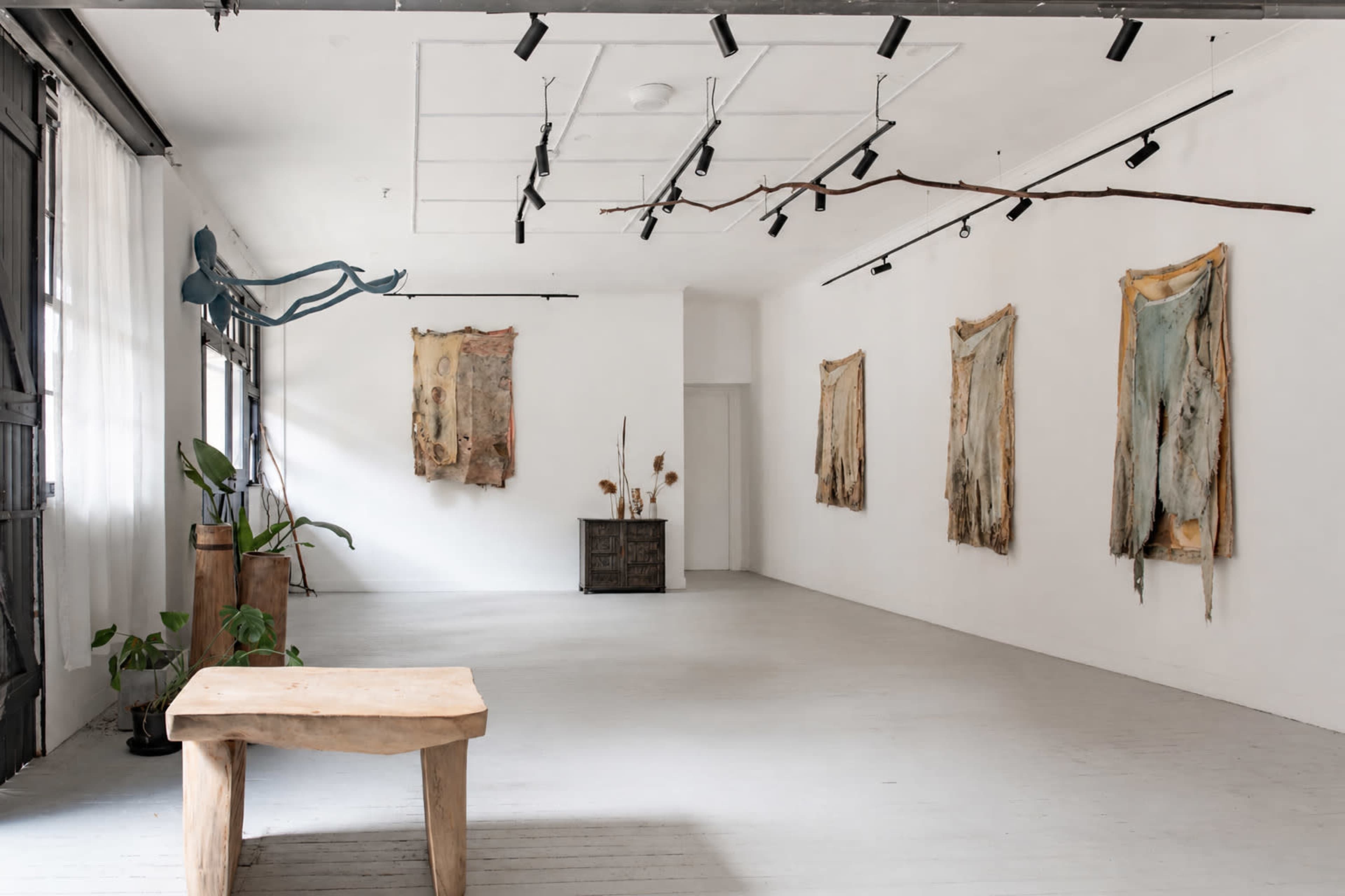 The image depicts an art gallery with several pieces of artwork mounted on white walls, a wooden table in the foreground, and minimal decor.