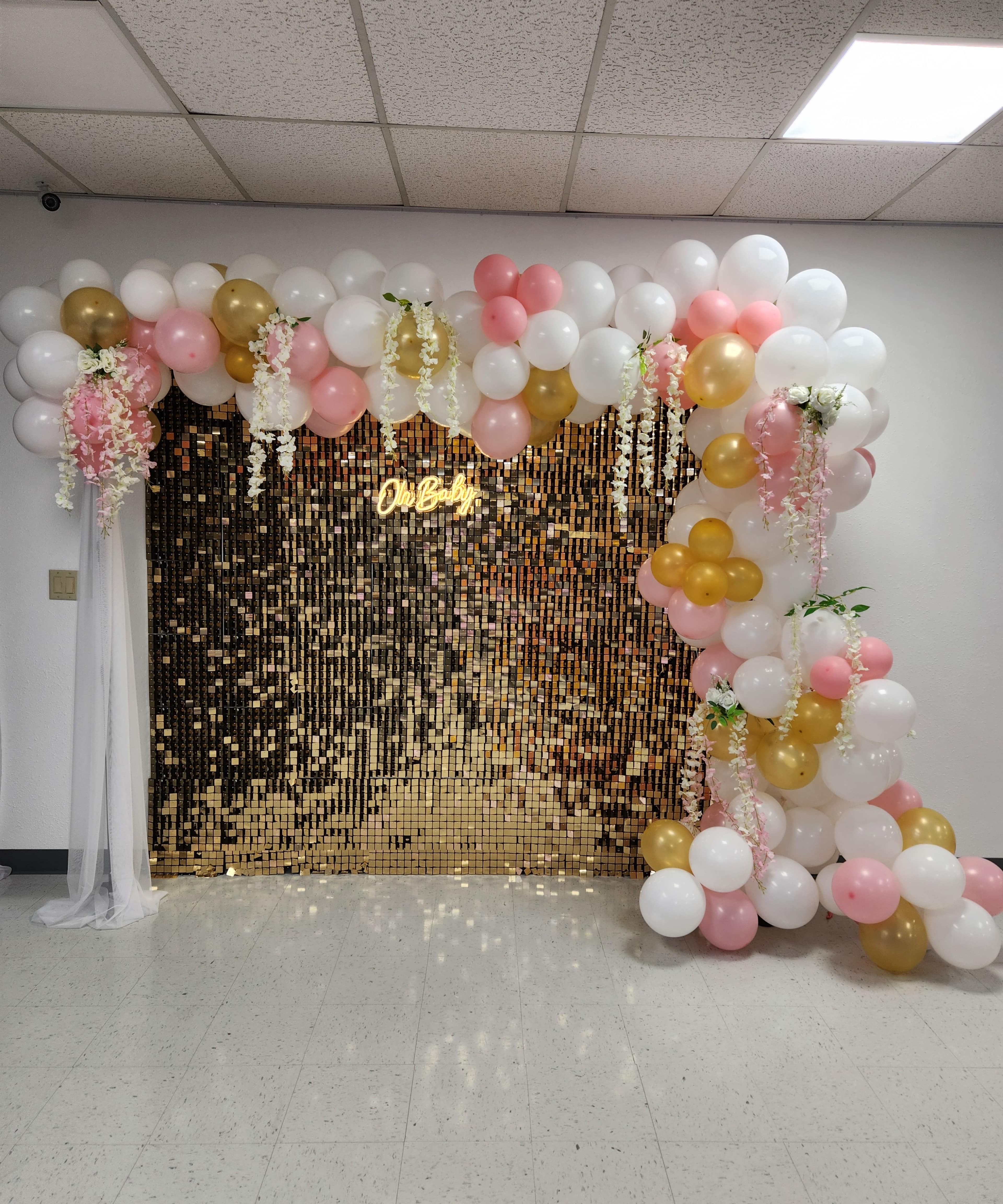 A decorated wall with a backdrop of golden sequins, adorned with pink, white, and gold balloons, and cascading floral elements.