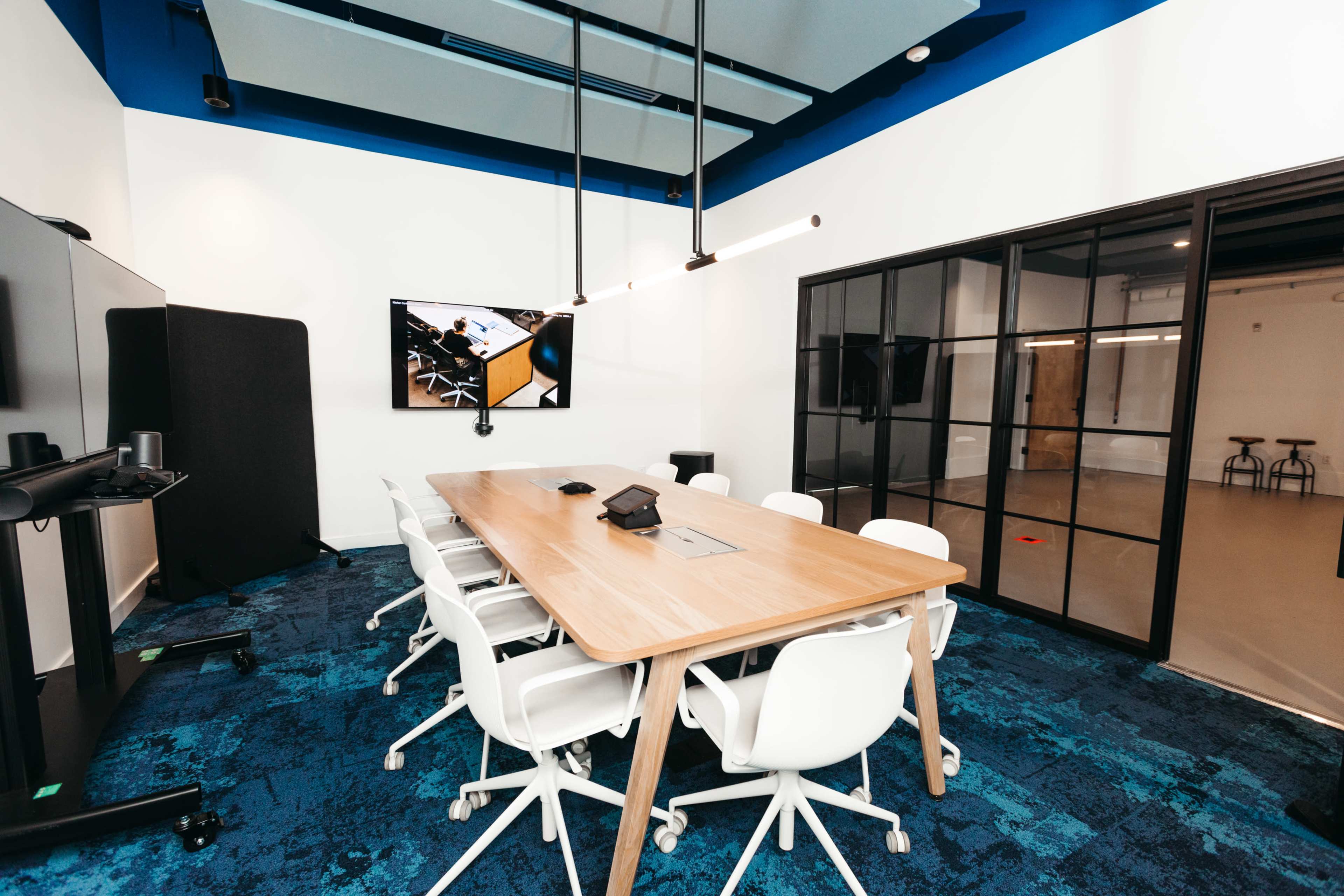 A modern conference room features a long wooden table surrounded by white chairs, with a large screen mounted on the wall and glass paneling.