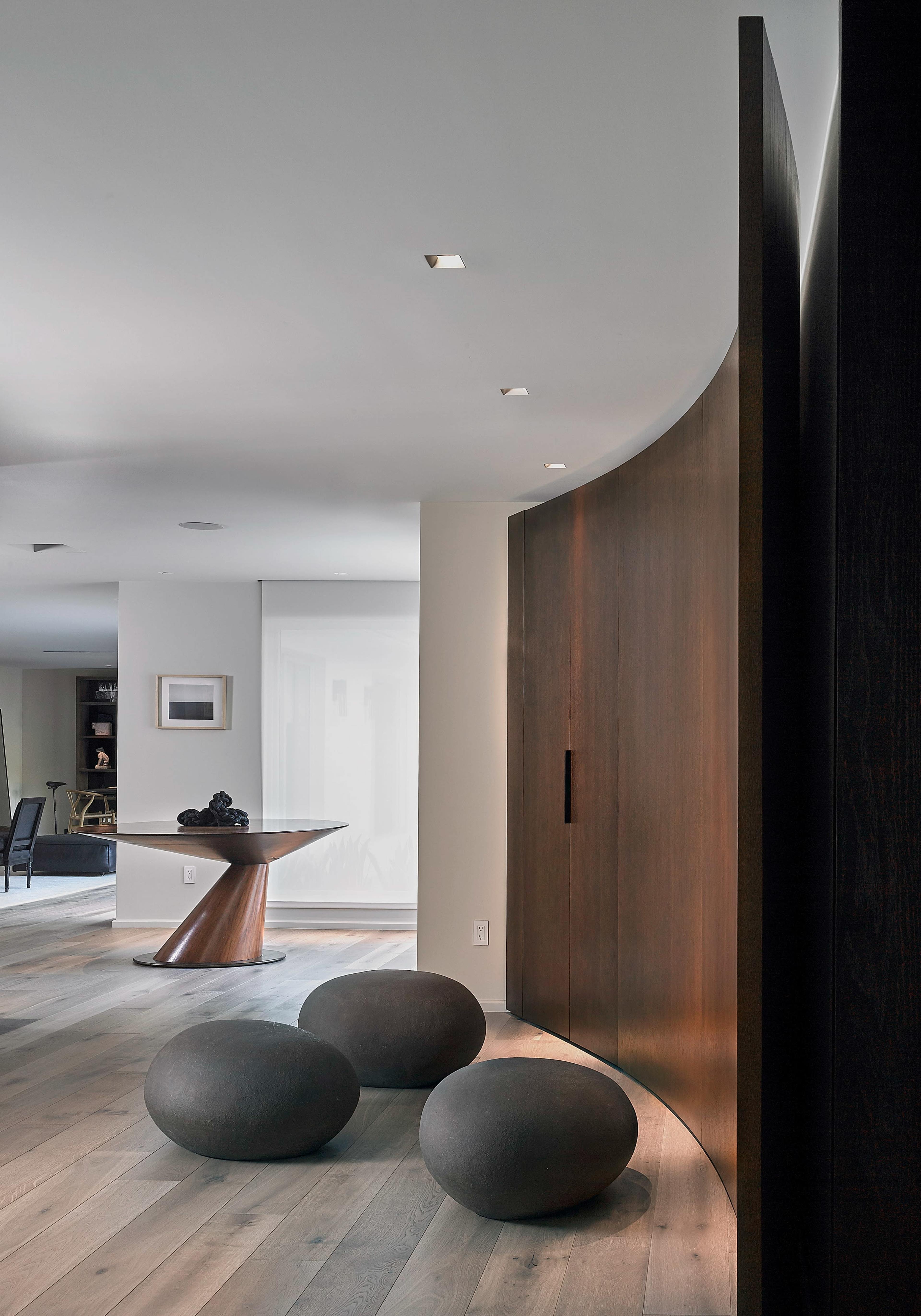 The image shows a modern interior with a curved wooden partition, a round table, and three spherical stone sculptures on a light wooden floor.