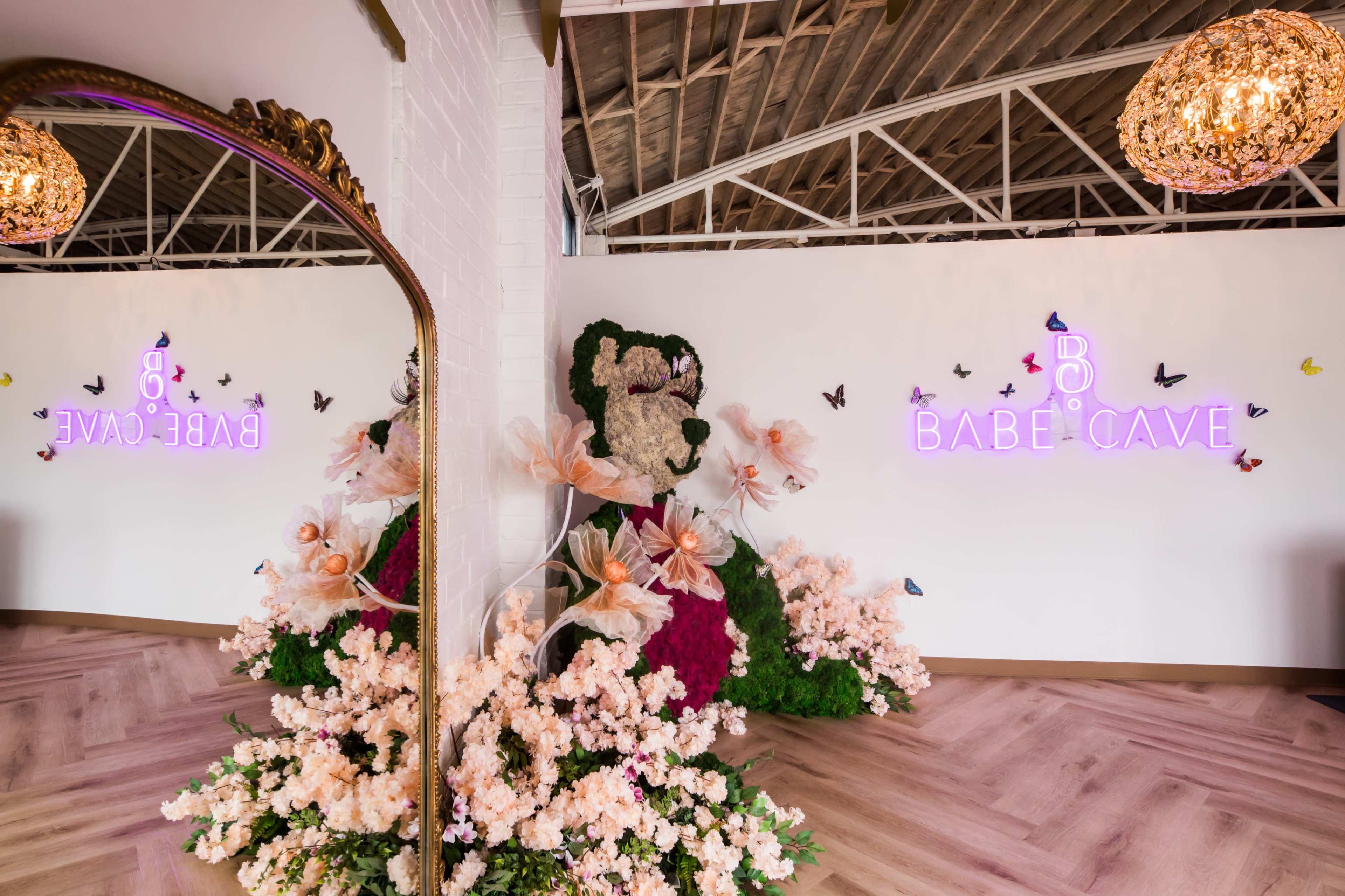 The image shows a vibrant interior of a space called "Babe Cave," featuring floral decorations, a large mirror, and decorative neon signage.