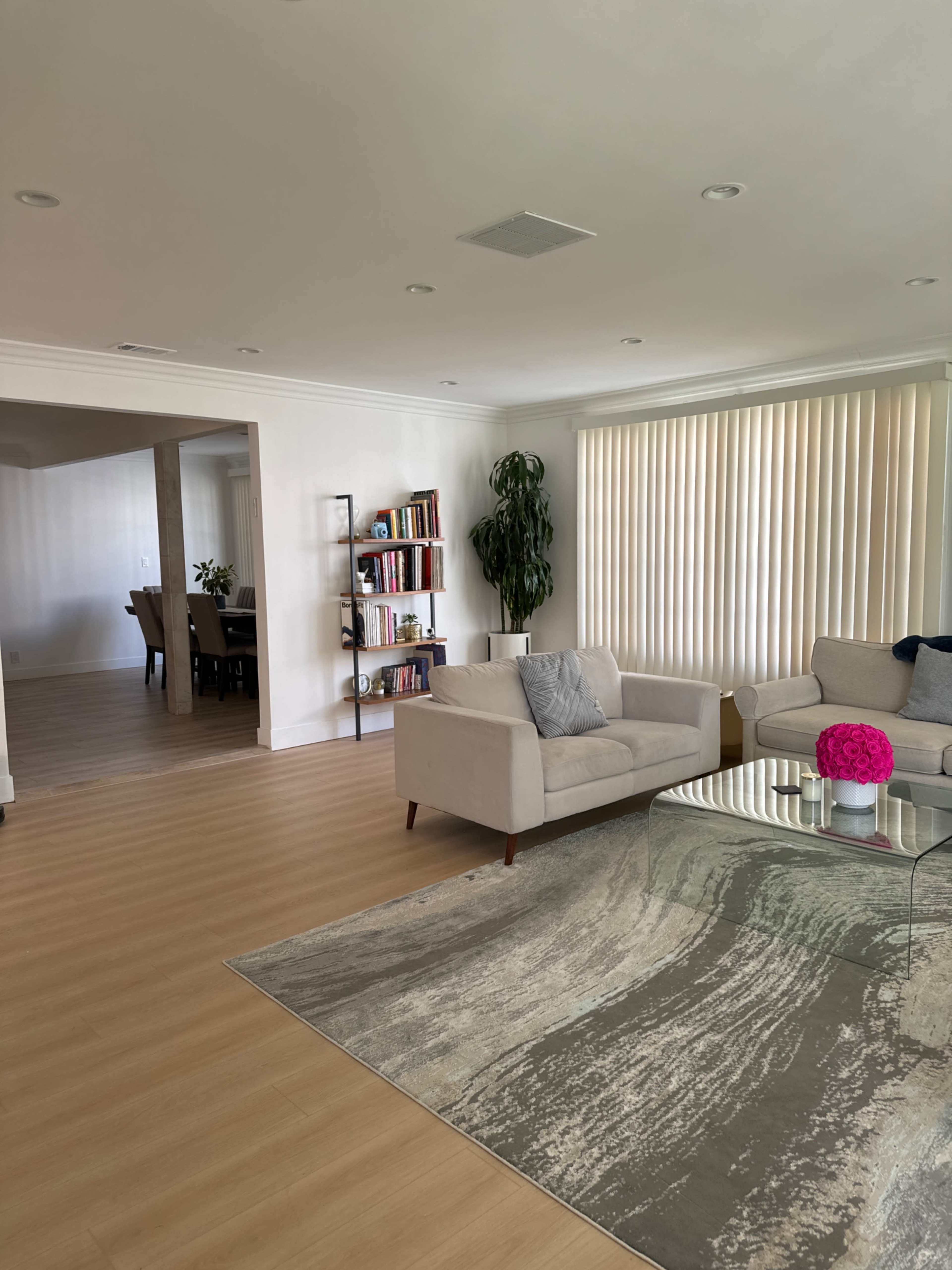 A modern living room with light wood flooring, a glass coffee table, a bookshelf filled with books, and large windows covered with vertical blinds.