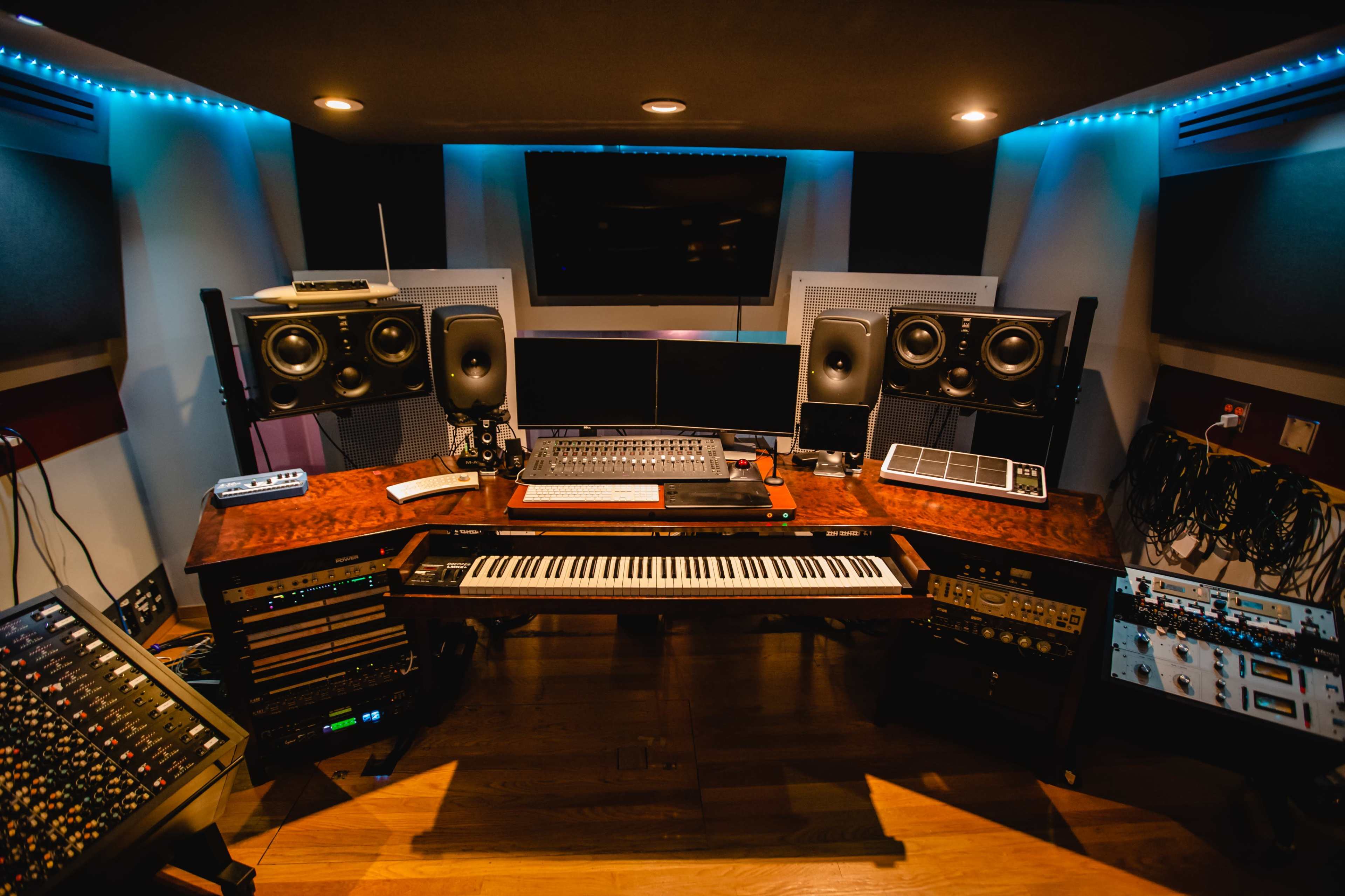 A music studio with a central wooden desk featuring a keyboard, multiple monitors, and audio equipment, surrounded by speakers and sound treatment panels.