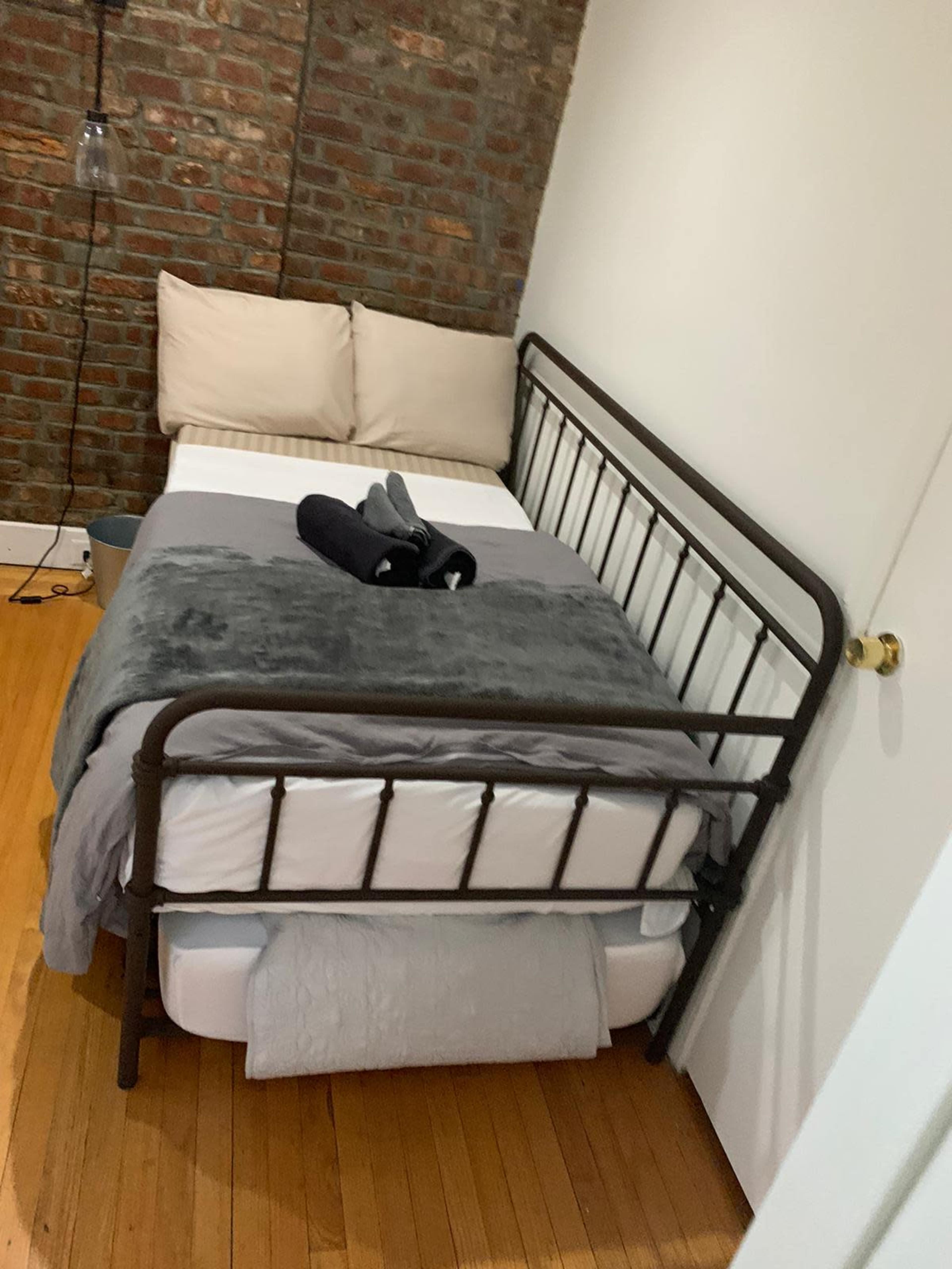 A metal-framed bed is placed against a wall with exposed brick, featuring gray bedding and neatly arranged towels.