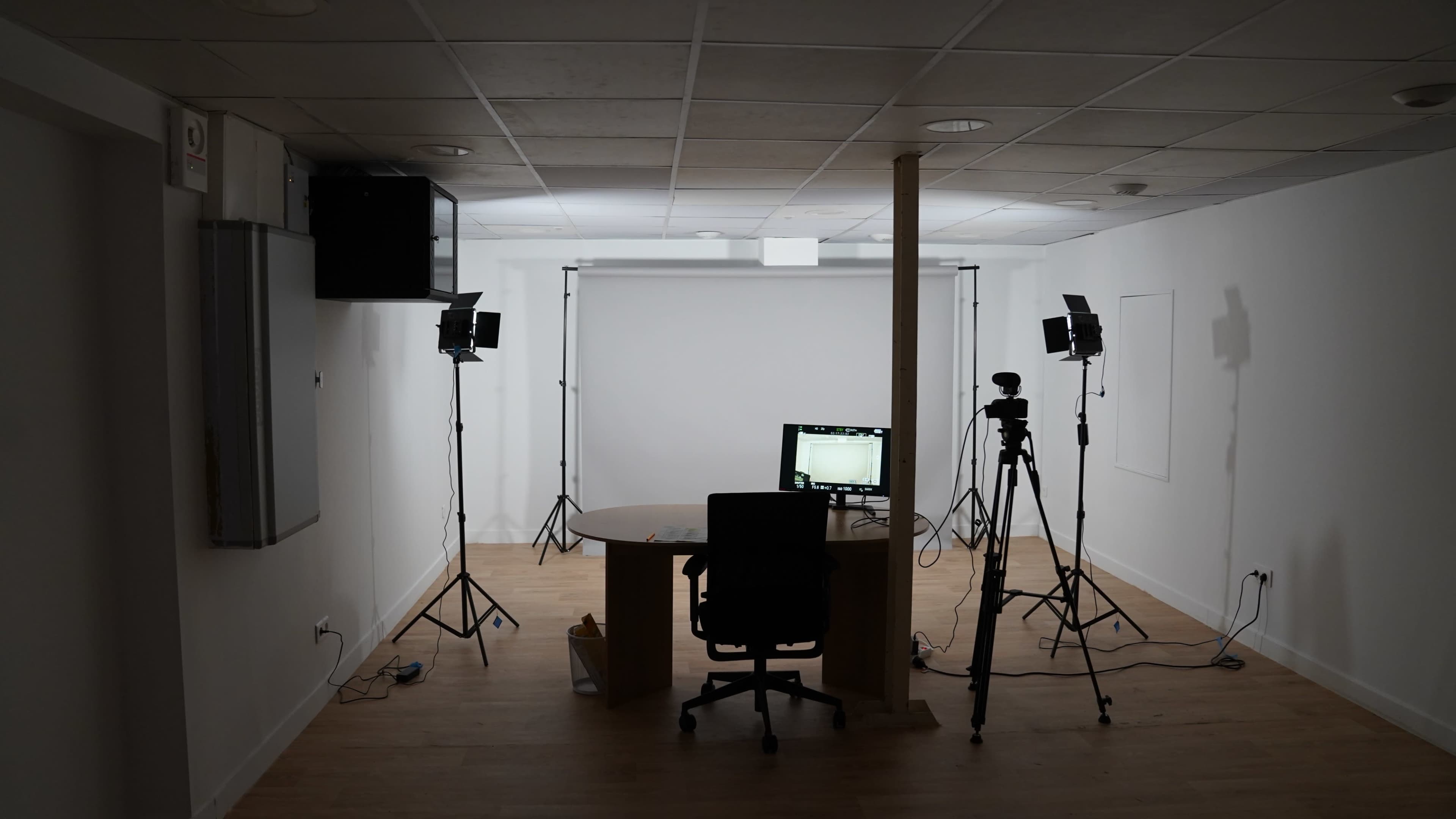 The image shows a sparsely furnished room with a desk, chair, and lighting equipment set up for video production.
