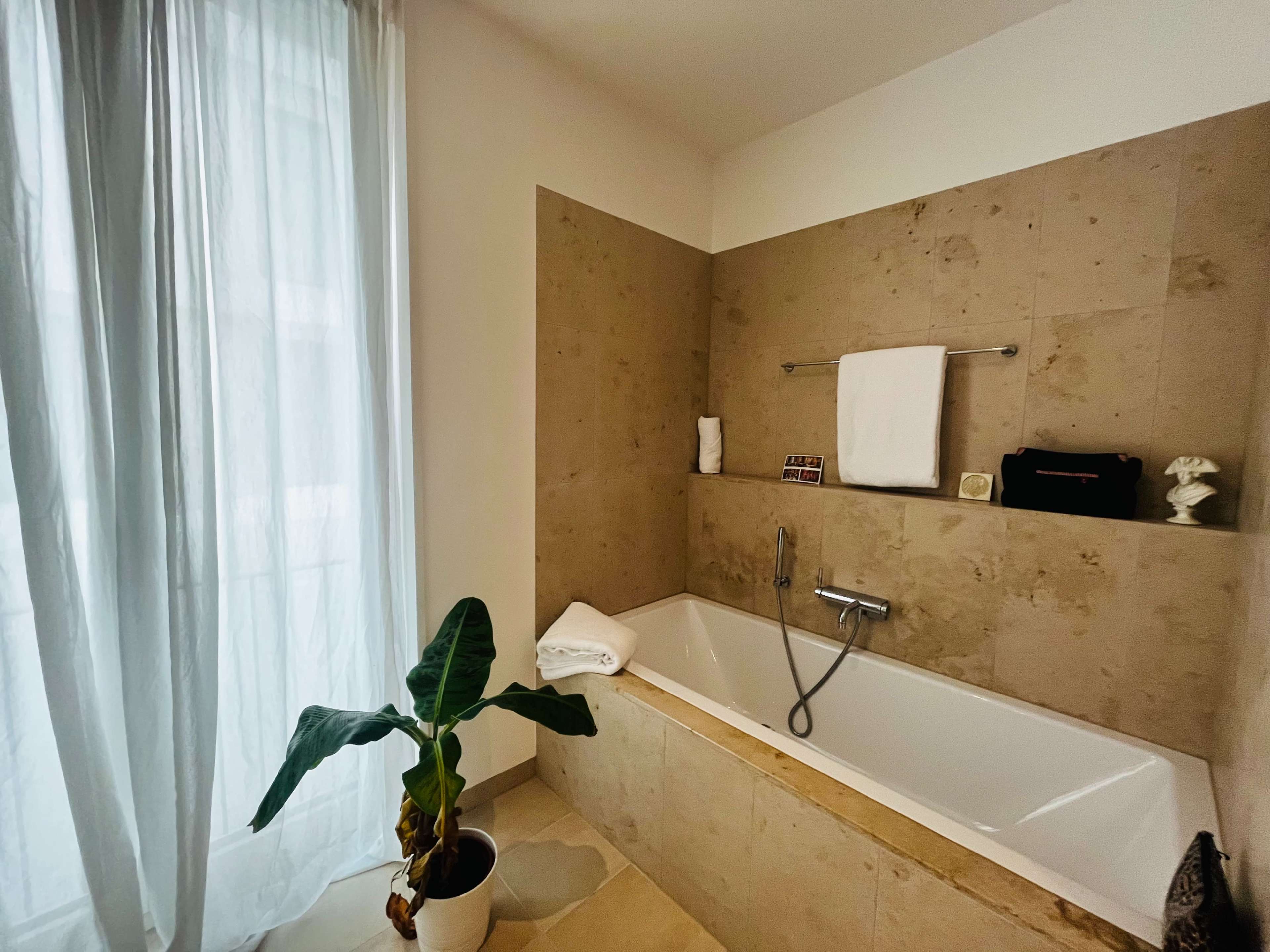A bathroom features a white bathtub surrounded by beige stone walls, with a window covered by sheer curtains and a potted plant nearby.