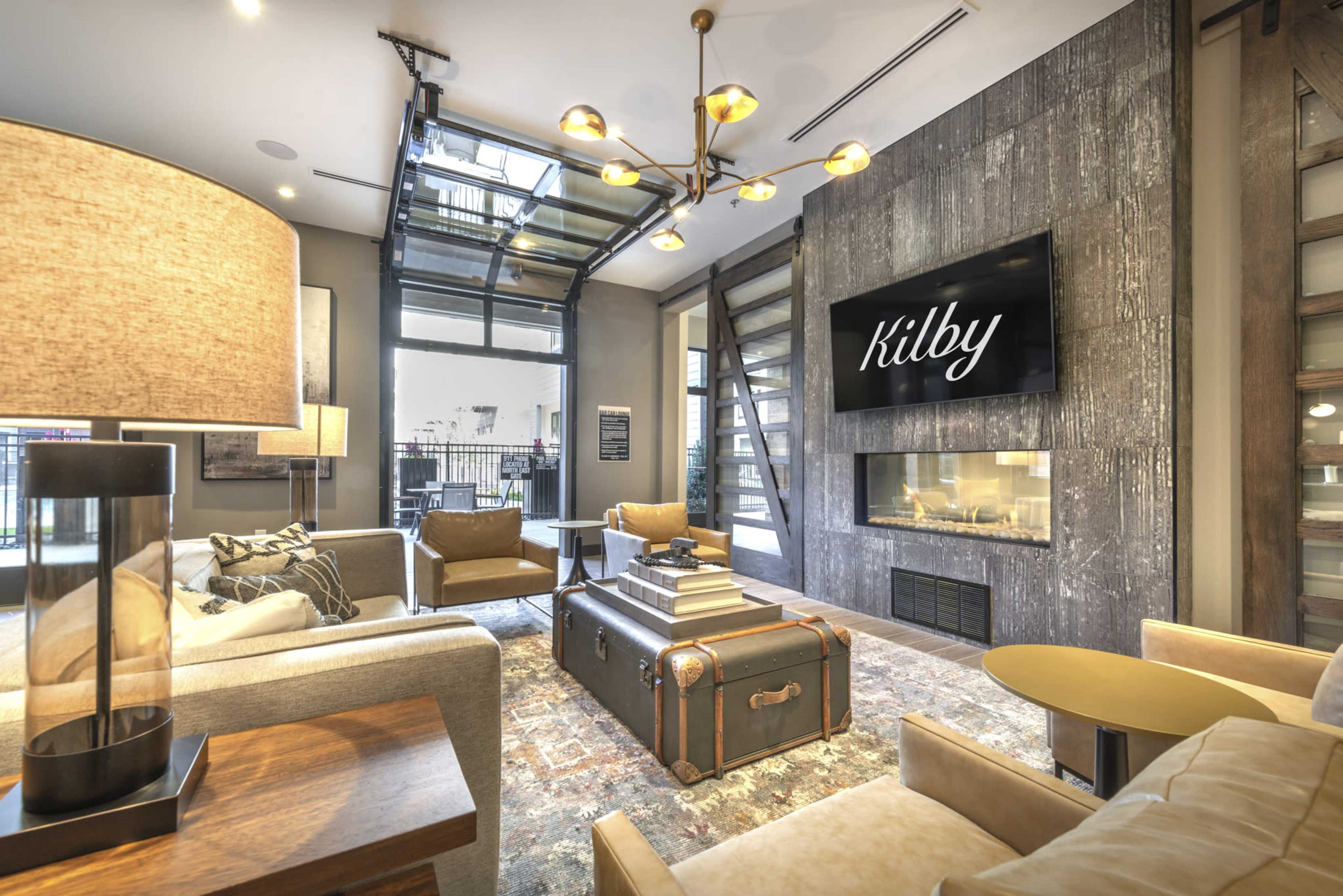 A modern lounge area with large windows, contemporary furniture, and a stylish fireplace featuring a screen displaying the name "Kilby."