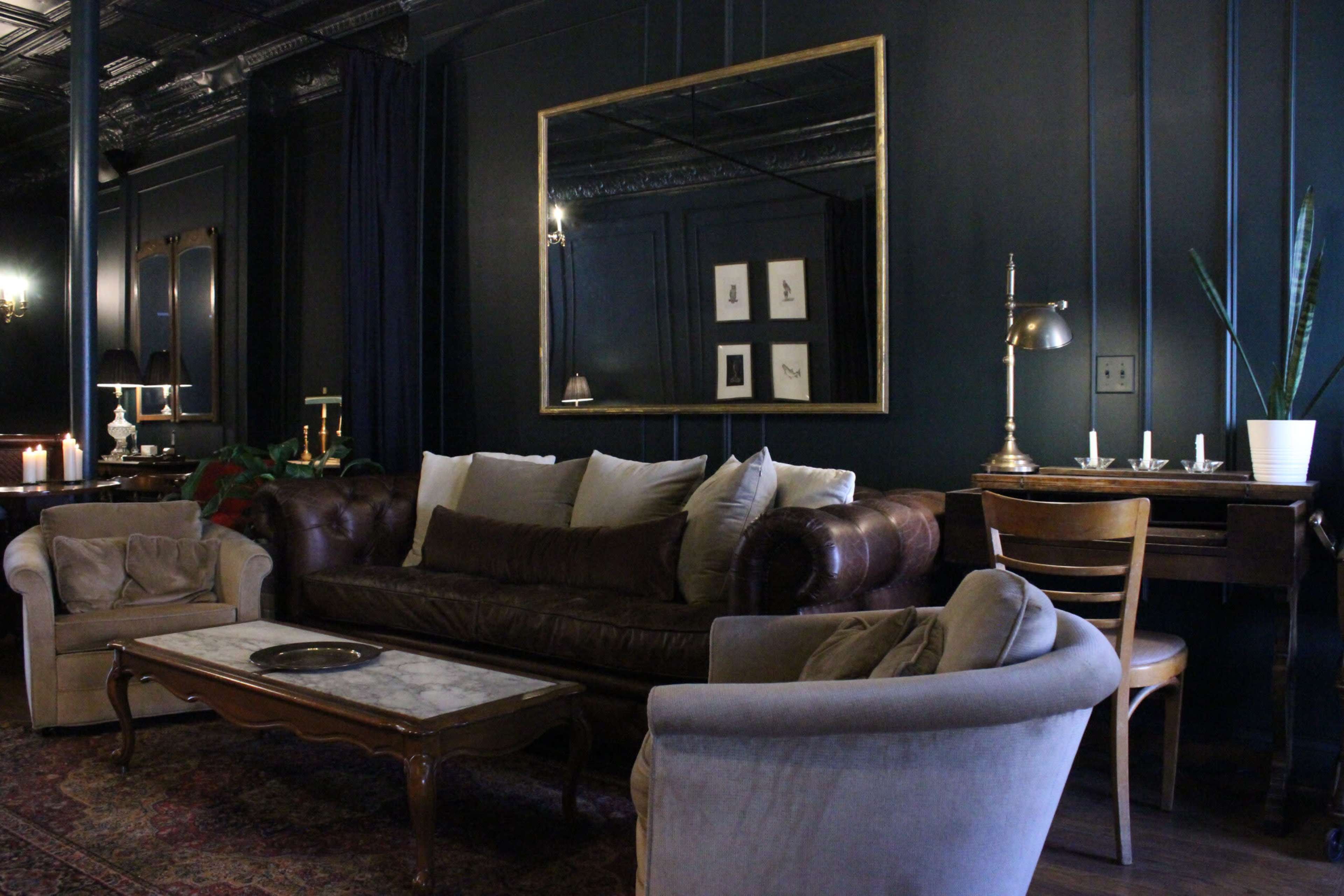 The image shows a darkly painted living room with a brown leather sofa, two armchairs, a marble coffee table, and a mirror on the wall.