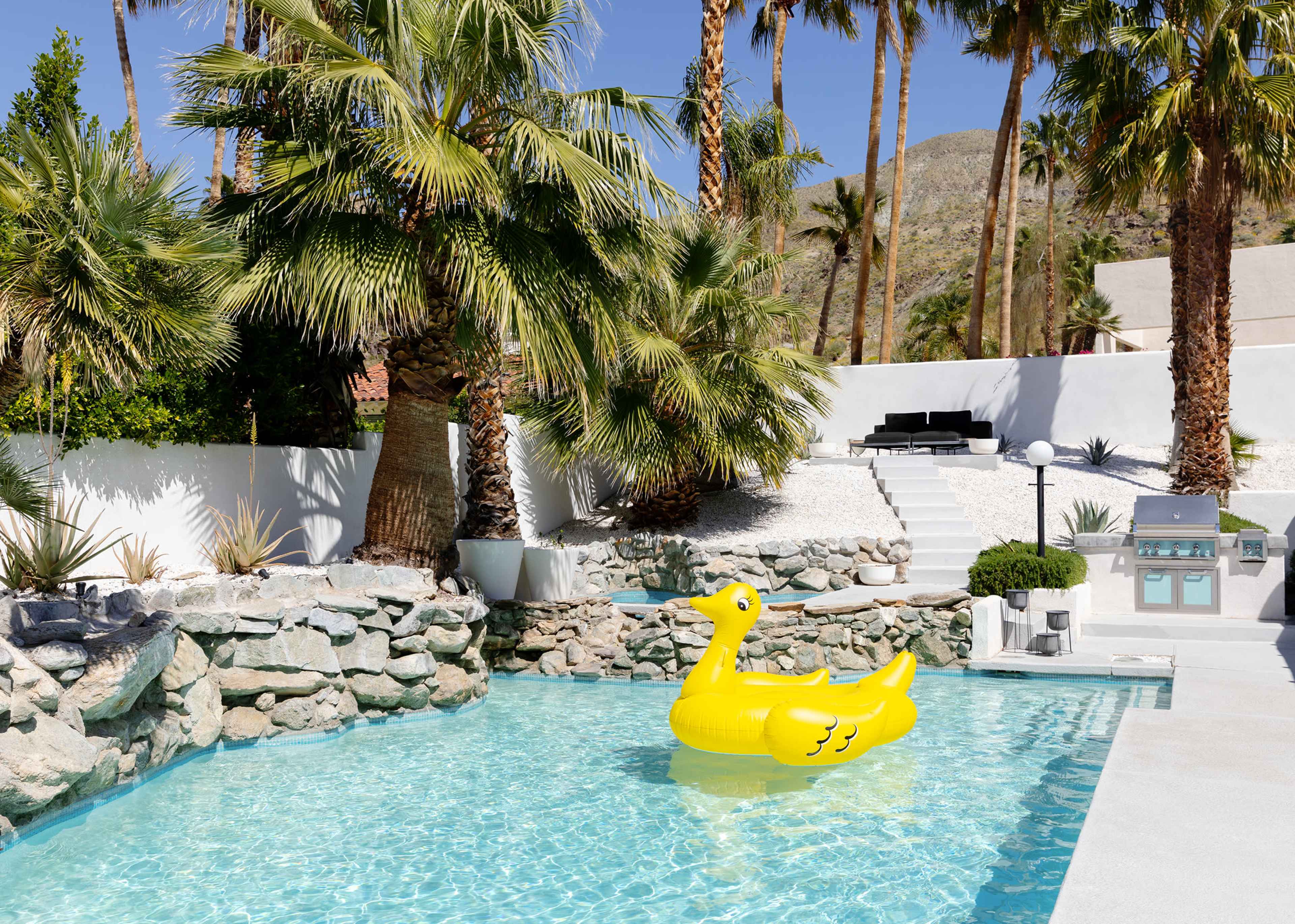 A large yellow duck-shaped float is positioned in a clear swimming pool surrounded by palm trees and stone landscaping.
