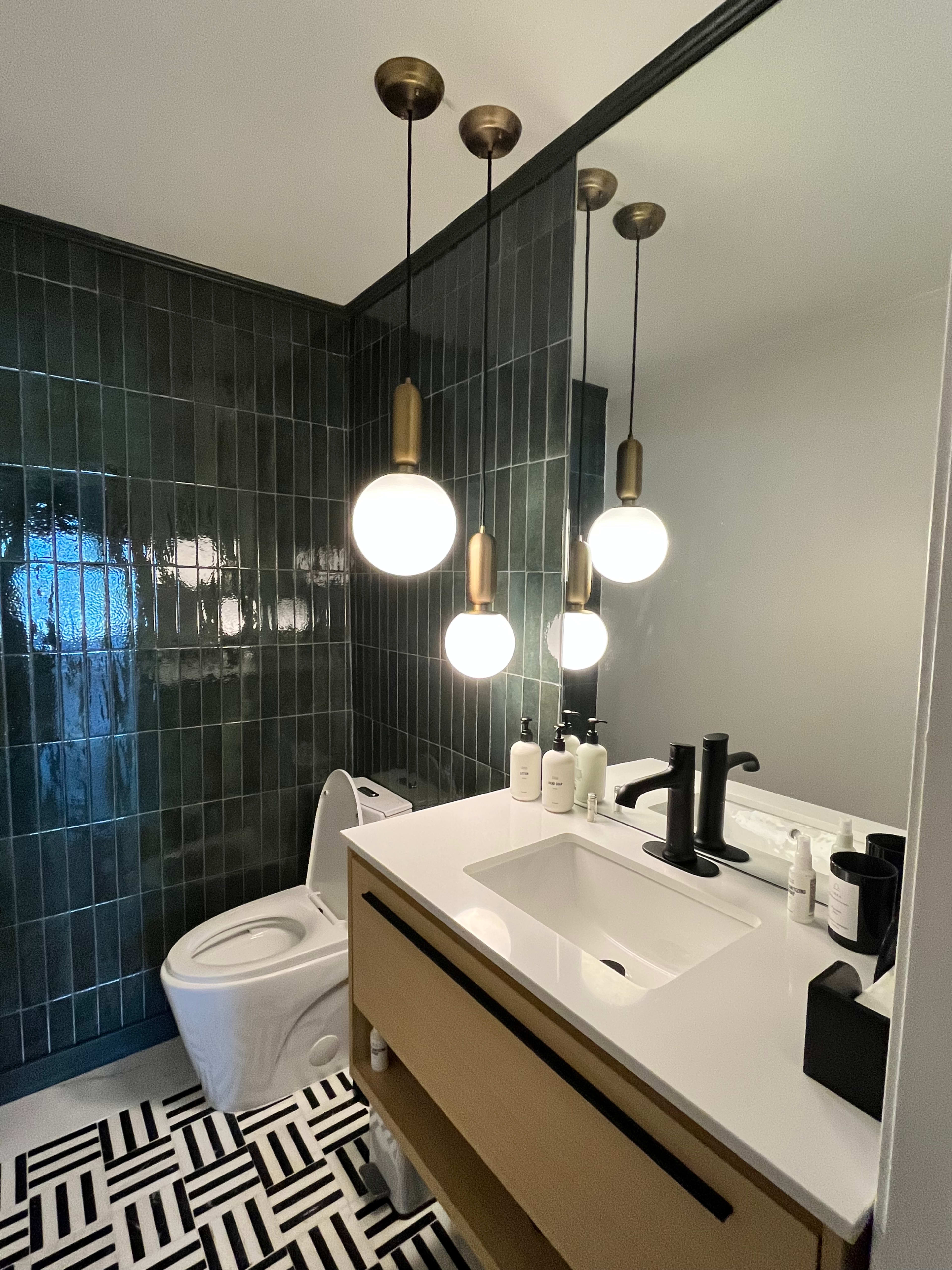 A modern bathroom with a white sink, wooden vanity, and three pendant lights above it, alongside a toilet and black-and-white patterned flooring.