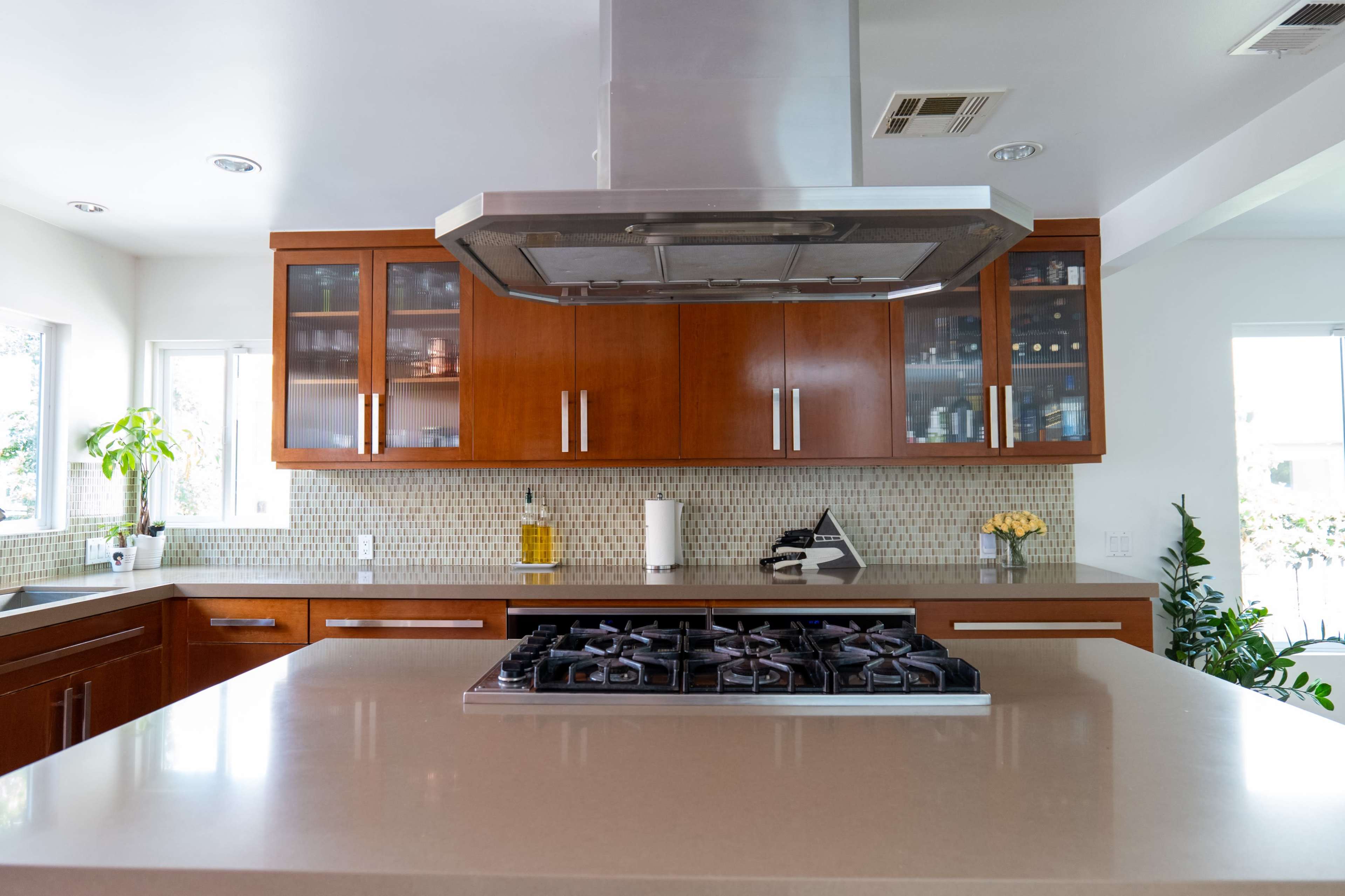 The image shows a modern kitchen with wooden cabinetry, a central island featuring a gas stove, and a silver range hood above it.