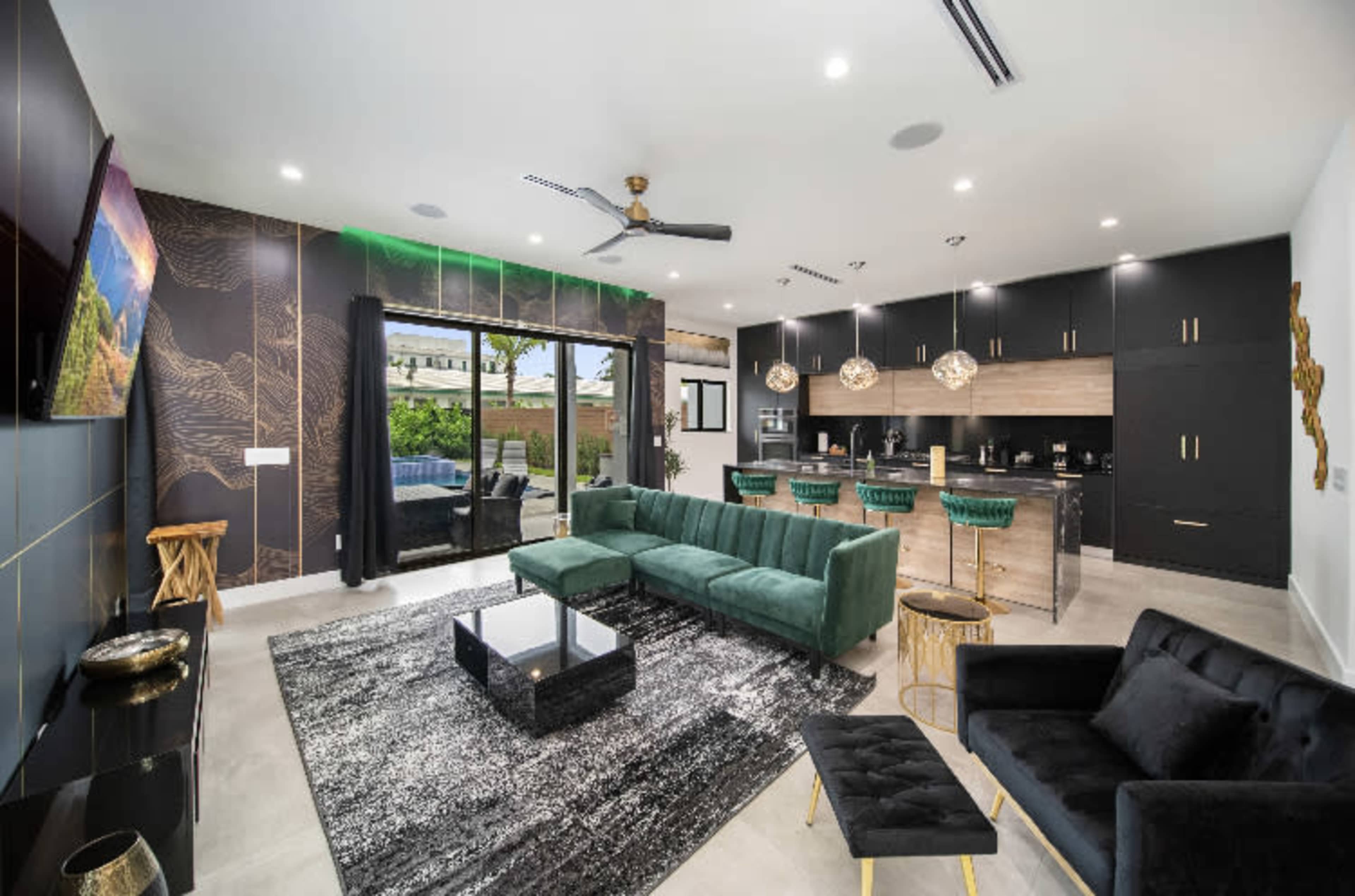 A modern living room features a green velvet sectional, a black coffee table, and a sleek kitchen with dark cabinetry and gold accents.