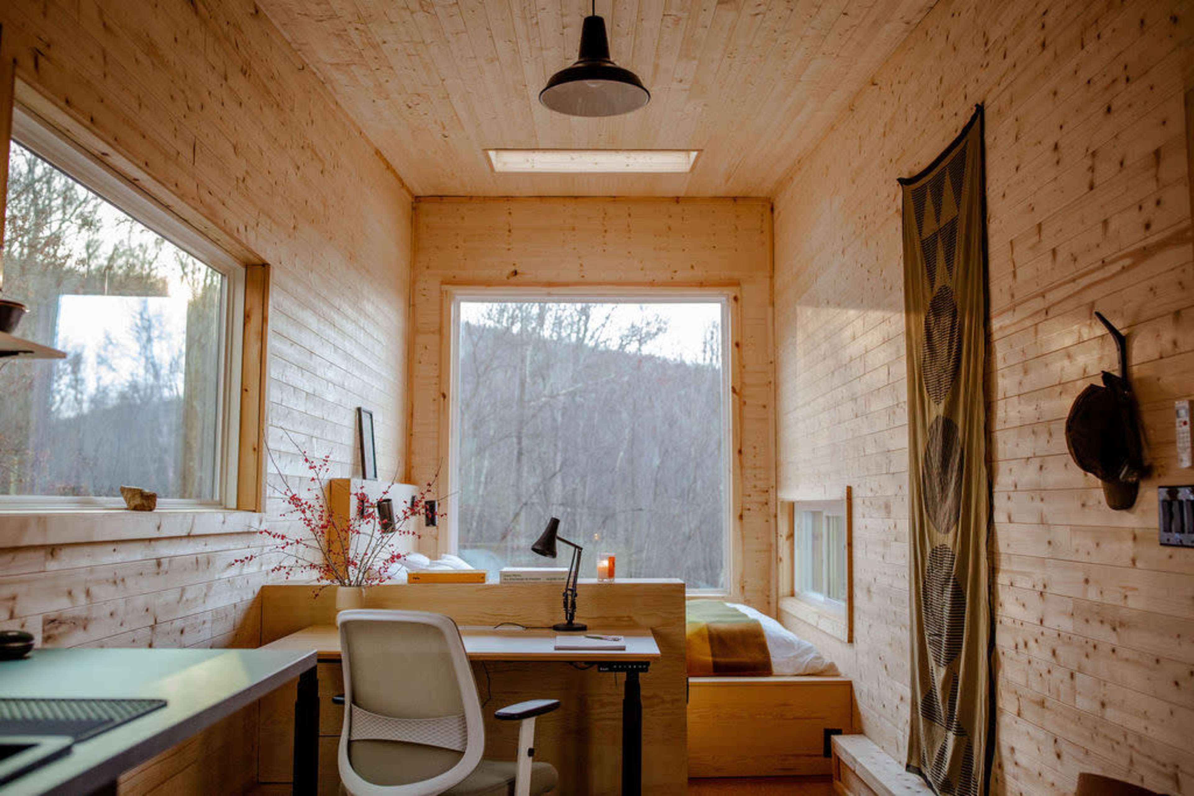 A minimalist wooden interior of a small room with a desk, chair, and a bed next to large windows that overlook a forested area.