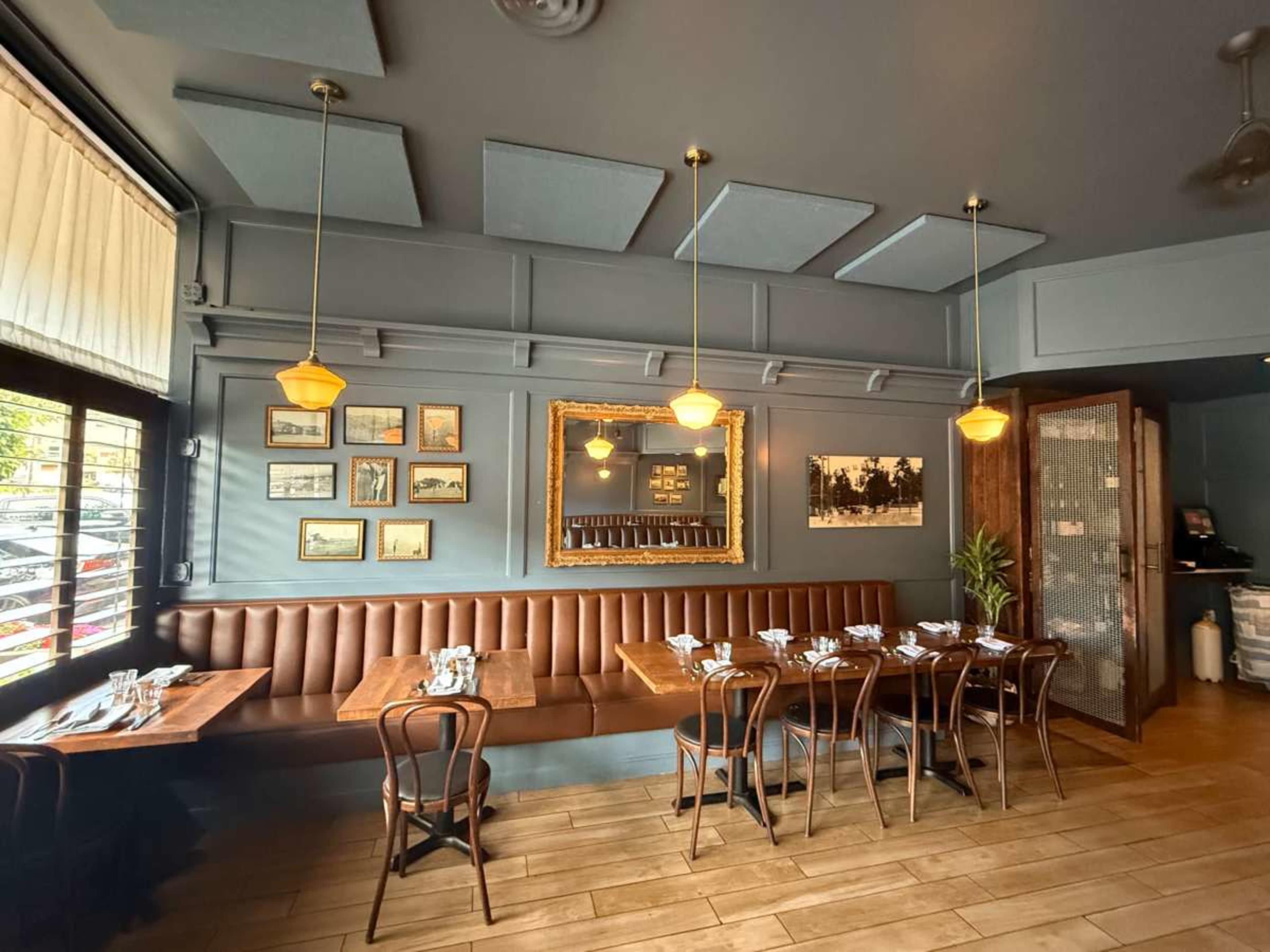 The image shows a restaurant interior with brown banquette seating, wooden tables set for dining, framed artwork on the walls, and pendant lighting overhead.