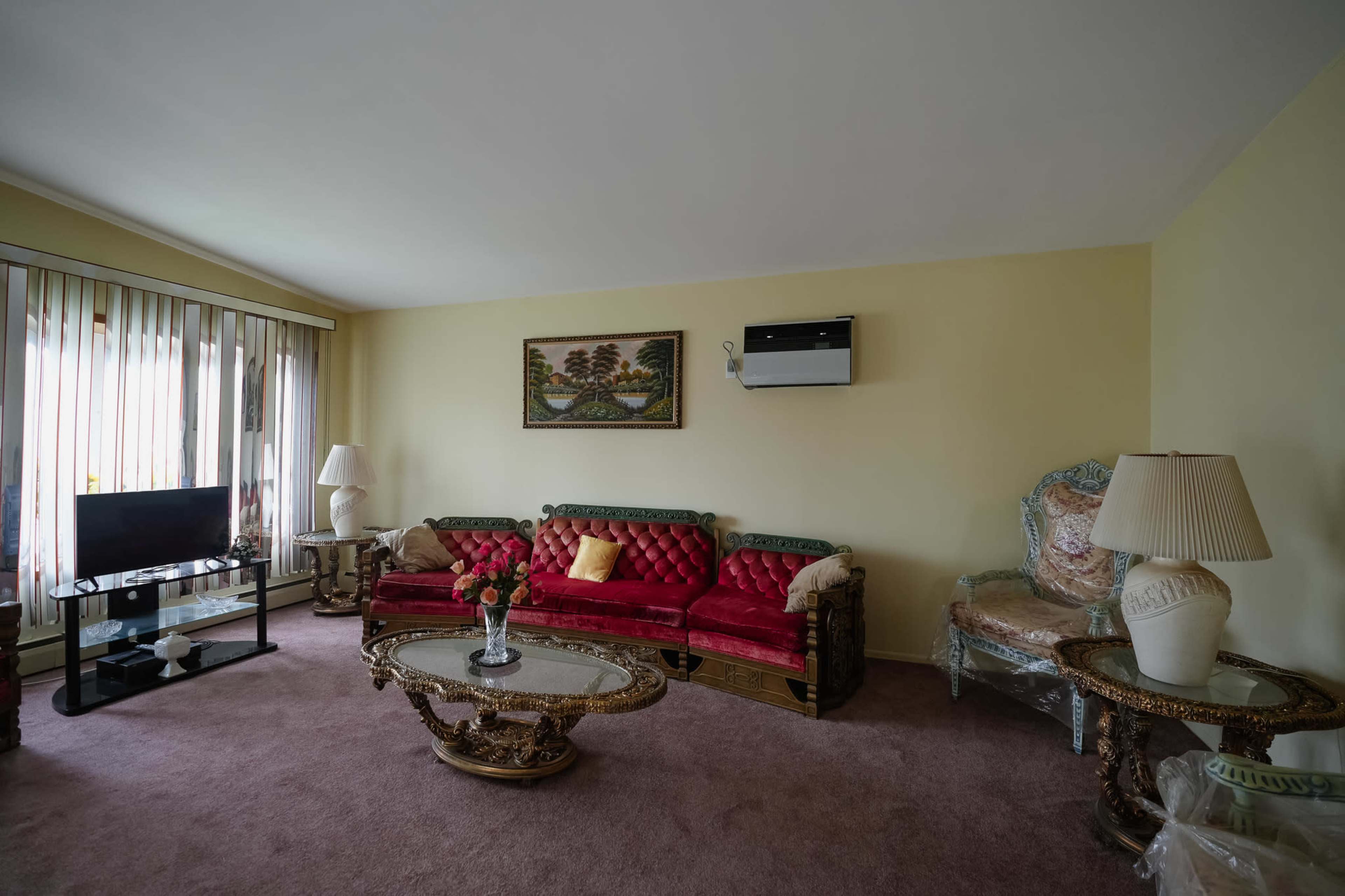 The living room features a red velvet sofa, an ornate coffee table, and a decorative armchair, with a television and large windows providing natural light.