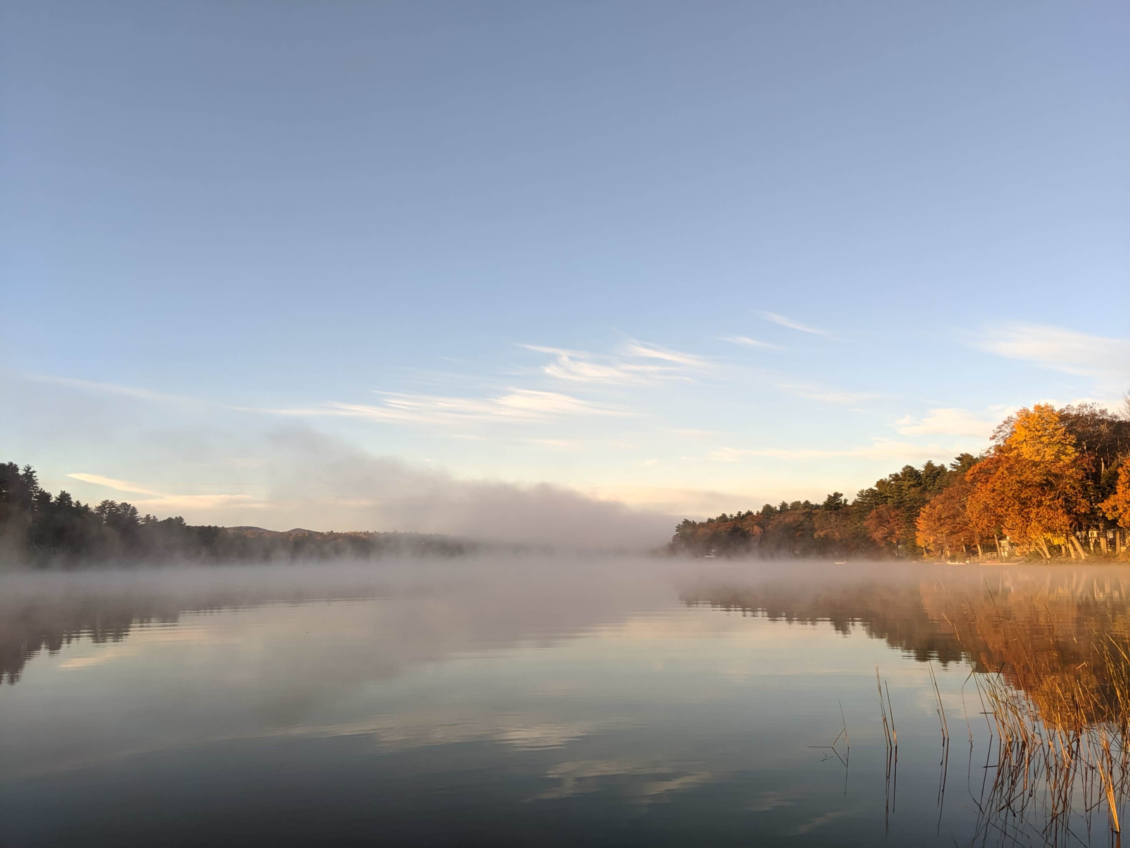 A calm lake reflects colorful autumn foliage and mist under a clear sky.