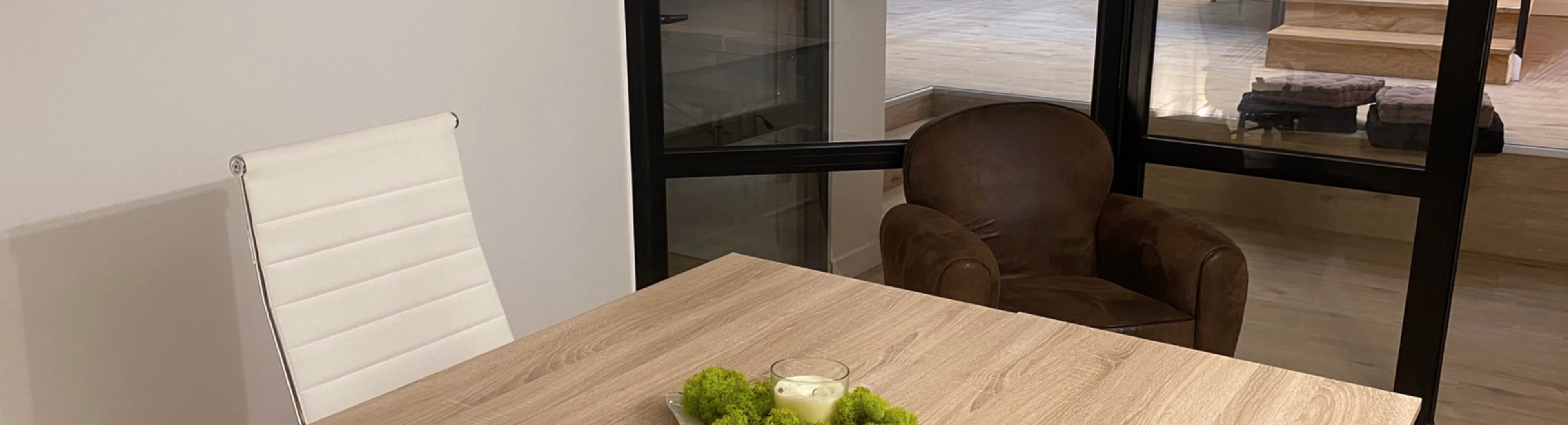 A wooden table with a bowl of green grapes sits in a bright room with a glass wall and a brown armchair nearby.